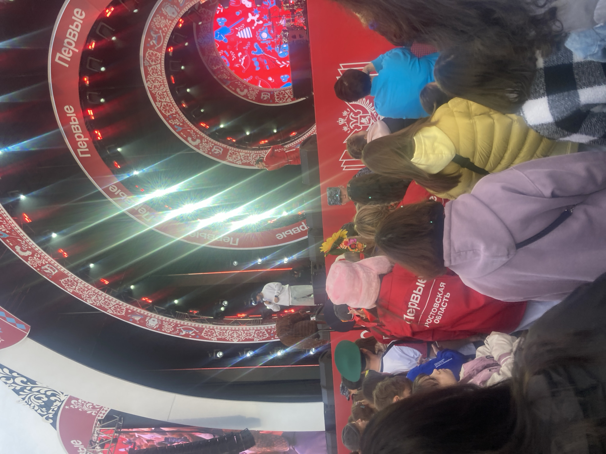 The image shows a large event taking place in Moscow, Russia. The backdrop features the word "Первые" (First) in Cyrillic, repeated around concentric circles. There is a stage with a red and white patterned background. In the foreground, there is a crowd of people. One person in a red jacket is wearing a jacket labeled "Первые РОСТОВСКАЯ ОБЛАСТЬ" (First ROSTOVSKAYA OBLAST). There is a person in white attire speaking on stage. The scene is brightly lit with stage lights.