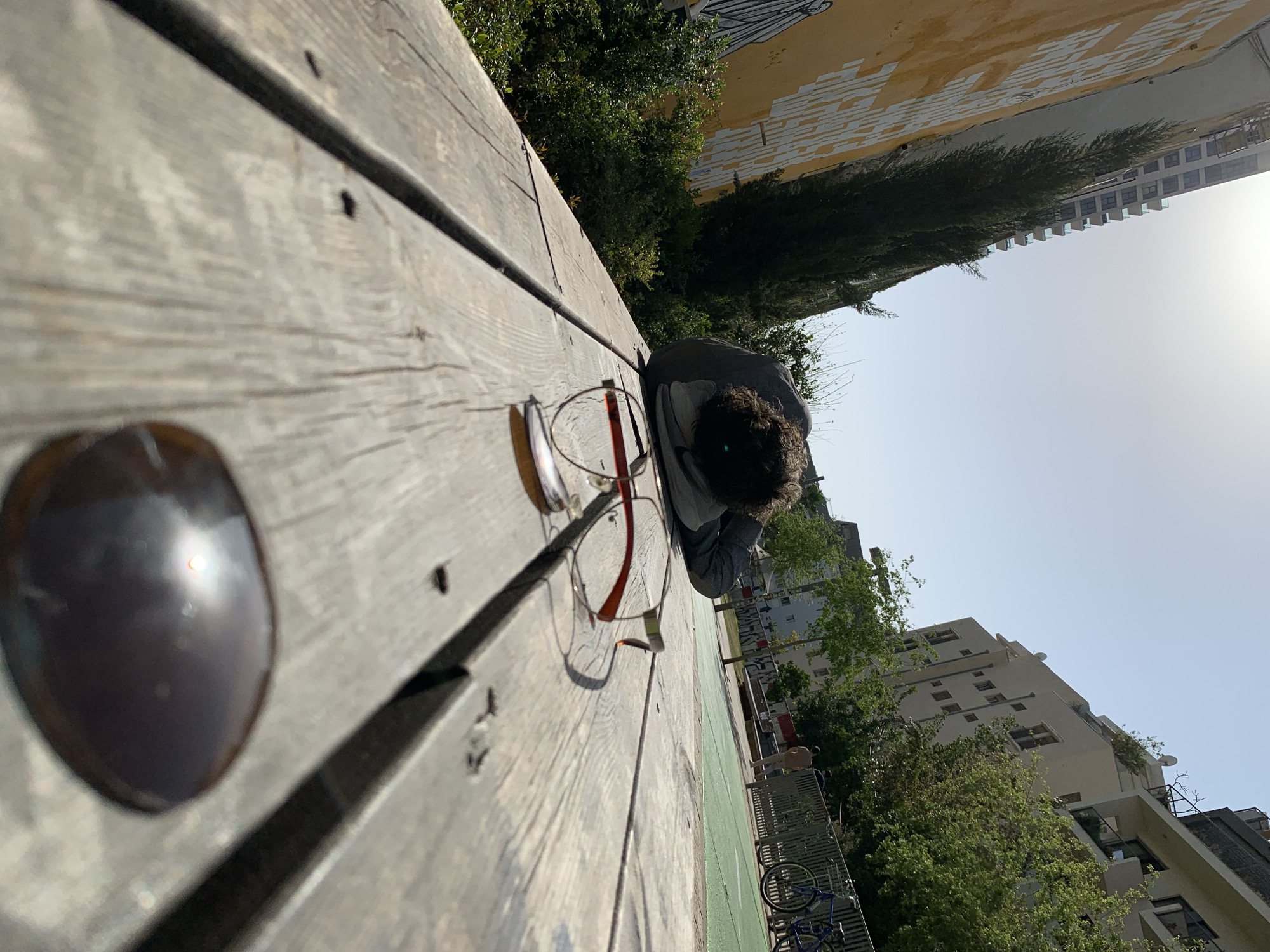 The image, originally oriented sideways, depicts an outdoor urban scene under bright daylight. In the immediate foreground, a light-colored wooden surface, possibly a picnic table or bench, extends diagonally across the frame. A blurry, dark, rounded object with a reflective highlight is prominent in the bottom left corner of this surface.
Further along the wooden surface, a person is seen lying face down, their head and upper torso visible. They have dark, curly hair and are wearing a grey top, appearing to be resting or sleeping. A pair of eyeglasses with reddish-brown frames is placed on the wood near them.
Behind the wooden surface, lush green foliage from trees and bushes creates a vibrant backdrop. Beyond the greenery, several buildings rise under a clear, bright sky. One building has a distinctive yellow facade covered with white, abstract markings or graffiti. Further in the background, a taller, modern-style building with multiple windows is visible. To the right of the resting person, a green paved area and a partially visible blue bicycle suggest a public park or recreational space, consistent with an urban setting in Tel-Aviv, Israel.
The strong sunlight and absence of clouds indicate a clear, sunny day, likely morning or afternoon. No decipherable text is clearly visible, only abstract patterns on the yellow building. The overall mood conveyed is one of quiet repose in a bustling city environment.