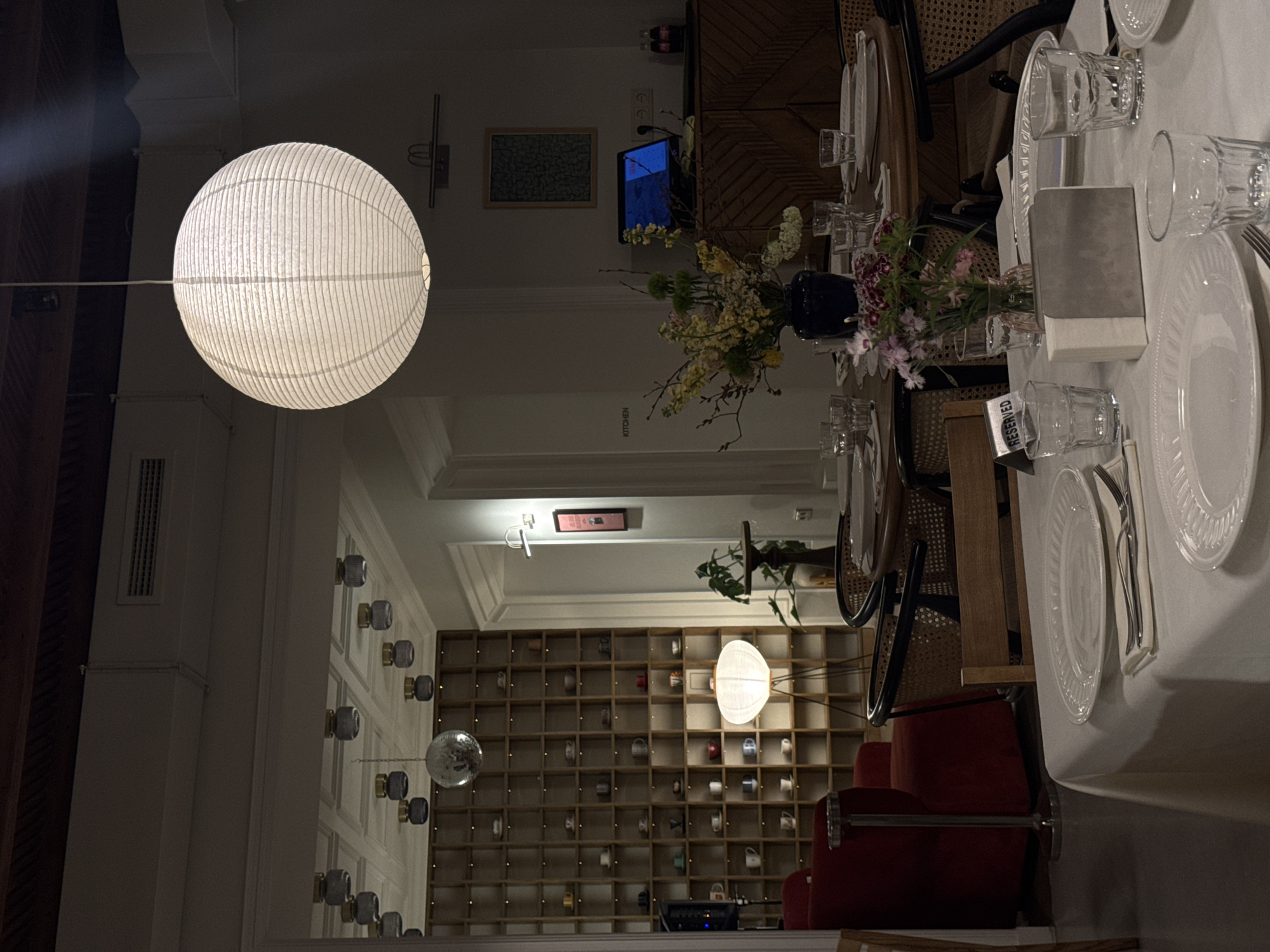 Captured indoors, the scene displays a high-angle perspective. At the forefront, a table setting is arranged with white plates, glassware, and silverware. A floral centerpiece adds a touch of nature to the setting. The dining arrangement suggests a formal or semi-formal event.
In the background, a cabinet is filled with items, indicative of a curated collection or display. A lamp casts a soft glow, contributing to the ambiance of the room.
A distinct structural element is observed: a ceiling installation featuring multiple light fixtures. This design element points towards an interior space designed with attention to detail.
Considering the setting and ambiance, the location may be a restaurant or a high-end dining establishment in Yerevan, Armenia. The detailed interior design elements and the curated collection in the cabinet further support this hypothesis.