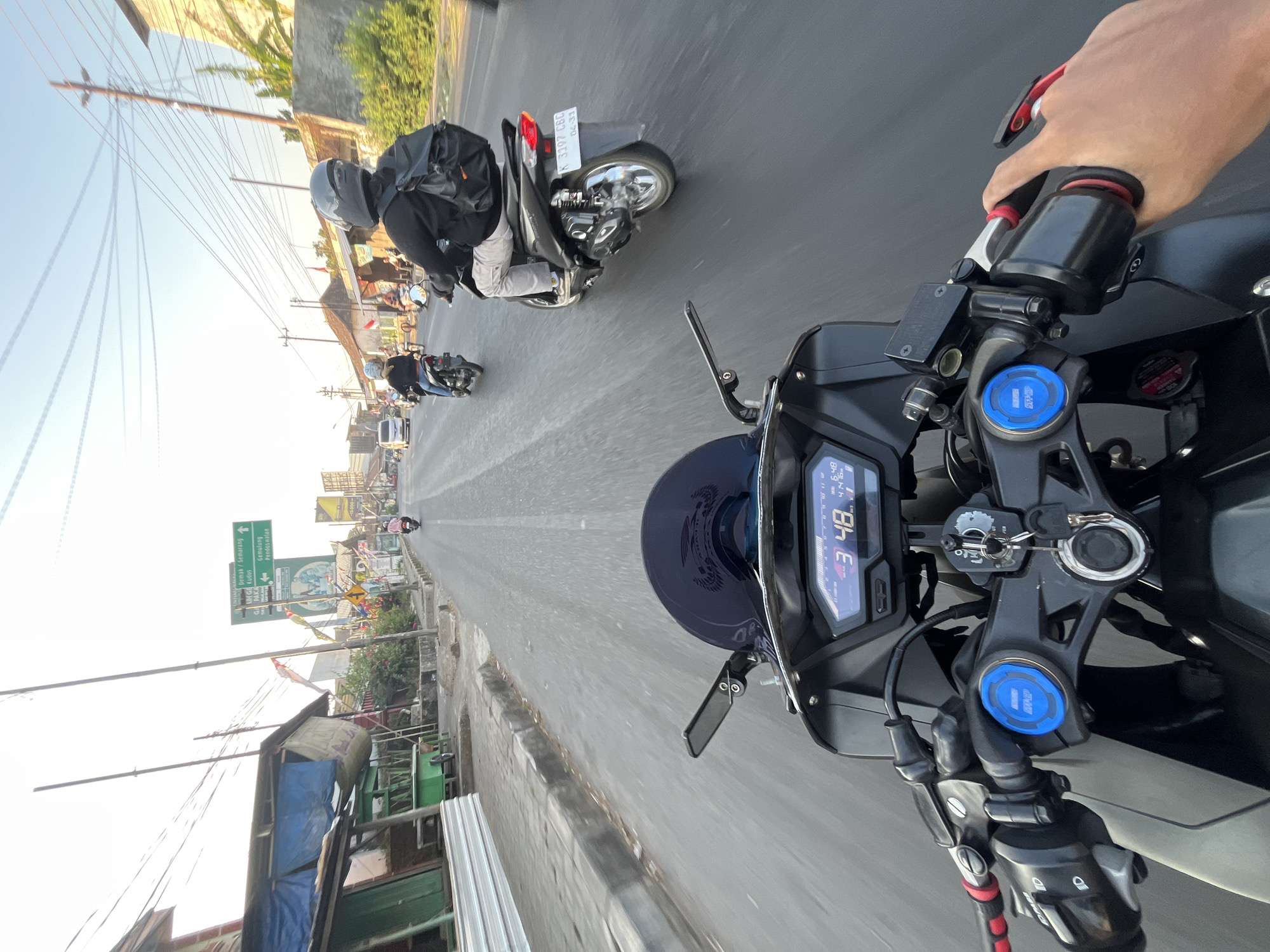 The image is a first-person perspective shot from a motorcycle traveling on a road in Kalinyamatan, Indonesia. The dashboard of the motorcycle displays a speed of "3 48". Several other motorcycles and a van are visible on the road, with riders wearing helmets. On the right side of the road, there are buildings and trees. On the left side, there are roadside stalls with blue tarps. A green directional sign is visible in the distance, indicating "Demak / Semarang," "Kudus," "Gajahmada," "Gemulung," and "Pekalongan." The sky is bright and clear, suggesting it is daytime.