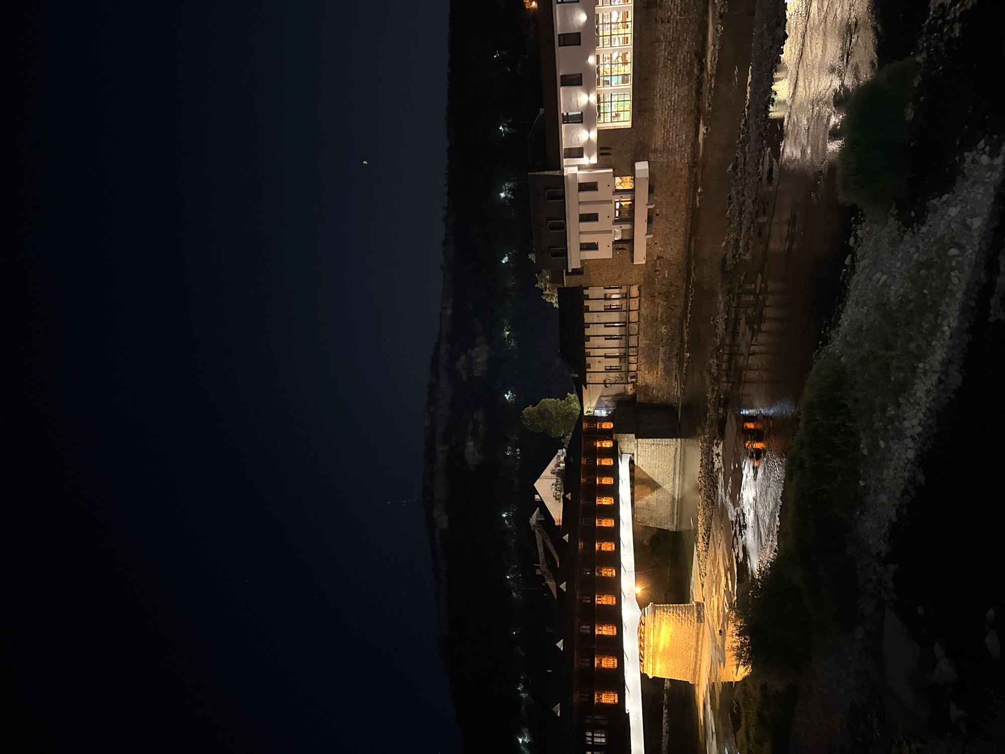 This is a nighttime exterior, medium-shot photograph of the covered bridge over the Osam River in Lovech, Bulgaria. The bridge, a covered wooden structure with numerous illuminated windows, is prominently featured spanning the dark water. On the far bank, several buildings with illuminated windows and facades line the river, reflecting onto the water's surface. The background consists of a dark, hilly landscape with some scattered lights and a visible radio tower or monument at the crest. The Osam River flows in the foreground, with its surface reflecting the lights from the bridge and buildings. The riverbanks are partially visible, featuring vegetation and rocks. The sky is dark and clear, with a faint crescent moon or planet visible.