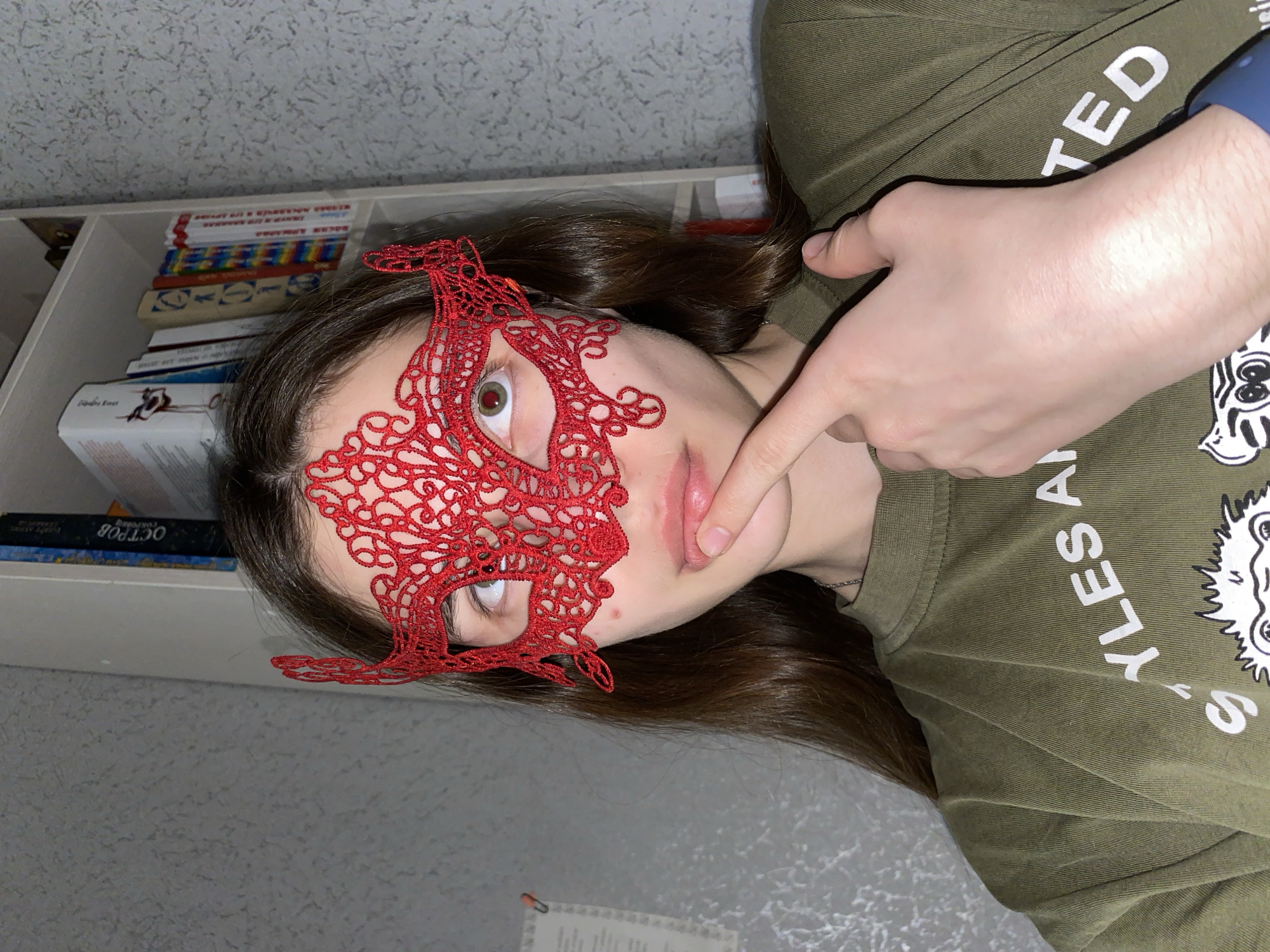 This indoor portrait features a young individual, likely in Votkinsk, Russia, posing with a red lace masquerade mask covering the upper part of their face. The person has long, dark brown hair and is wearing an olive-green t-shirt. Their visible left eye, seen through the mask's intricate pattern, appears wide and has a reddish tint in the pupil, indicating the use of a flash.
A fair-skinned hand, possibly belonging to the subject or another person, is positioned near the subject's mouth, with the index finger touching the lower lip. This hand also sports a blue wristband, potentially a smartwatch. The olive-green t-shirt has white text, partially visible and oriented sideways or upside down, reading "TED" and "YLES S" among other letters.
The background shows a light-colored, textured wall and a white bookshelf filled with books. Some books have colorful, possibly rainbow-striped, spines, while others display Cyrillic text. One book spine near the bottom left clearly shows "ОСТРОВ" (OSTROV, meaning "Island"). The lighting suggests an interior setting, possibly evening, with artificial light contributing to the red-eye effect. The overall mood of the image is one of playful mystery or anonymity due to the mask.