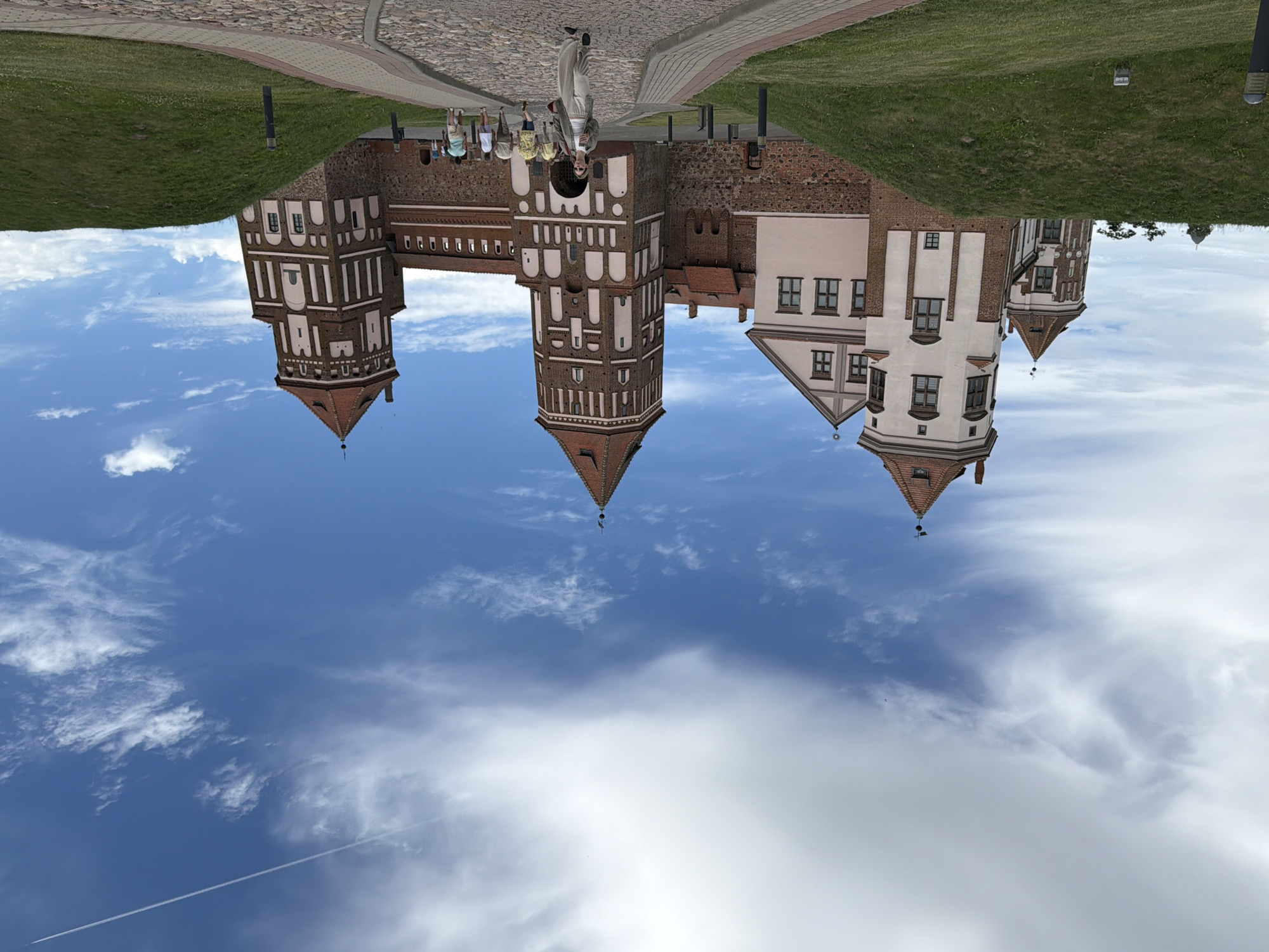 The image shows a castle with several towers and parts of its walls. The castle is made of red brick and has white accents on the towers. There are several people walking on a cobblestone path in front of the castle. The castle is surrounded by green grass and a bright blue sky with some white clouds. One of the towers has a red conical roof. The image is taken from a low angle, looking up at the castle. The sun appears to be shining, and the weather seems pleasant. The scene evokes a sense of historical grandeur and tourism. There is no visible text in the image.