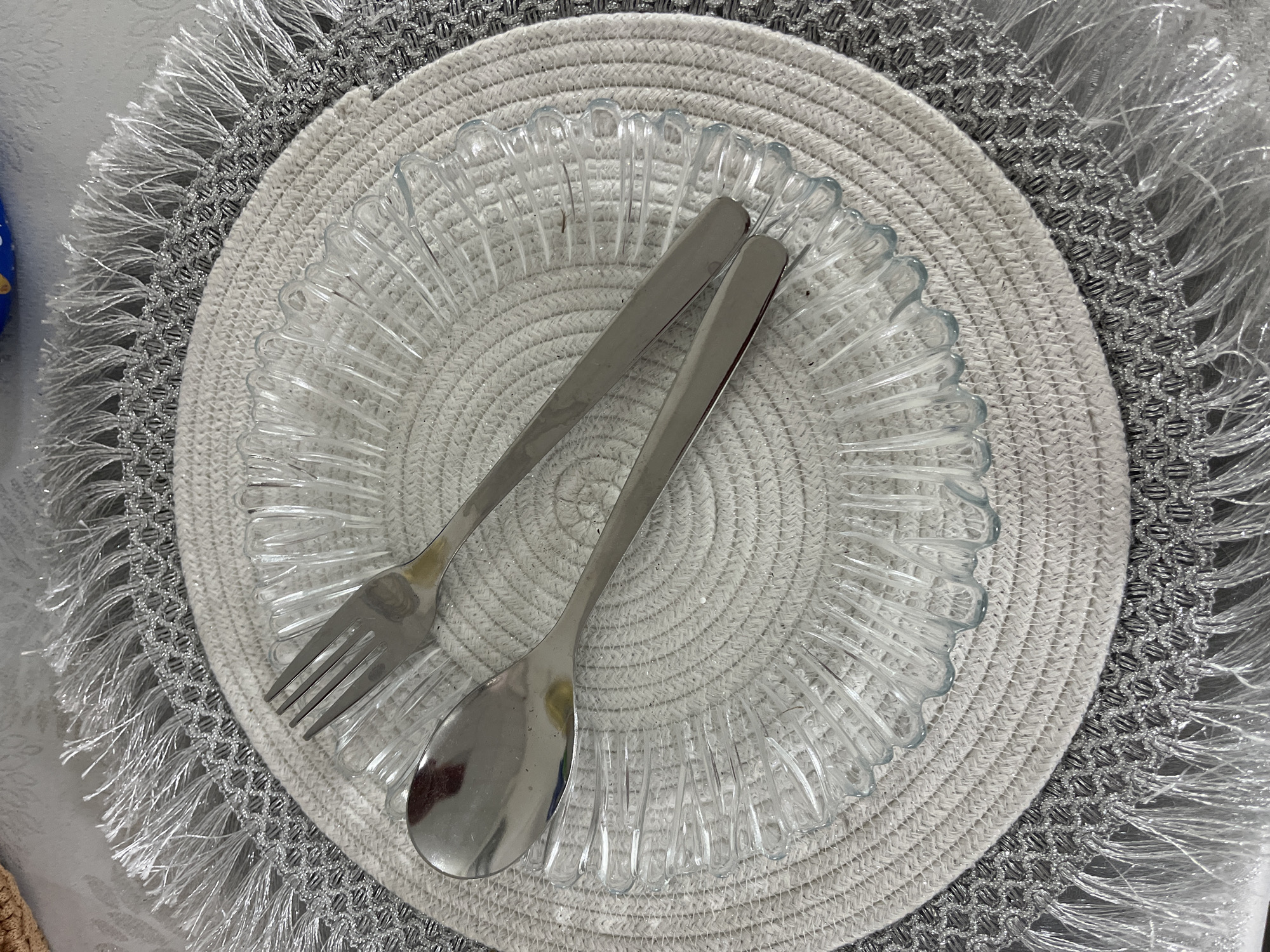 A silver spoon and fork are placed on a clear glass plate. The plate is on a round, woven placemat with a decorative silver fringe. The placemat is on a grey surface, possibly a table. There are no people or animals in the image, and no discernible location in the city of Hrodna, Belarus. The scene appears to be a still life arrangement of dining utensils and placeware.