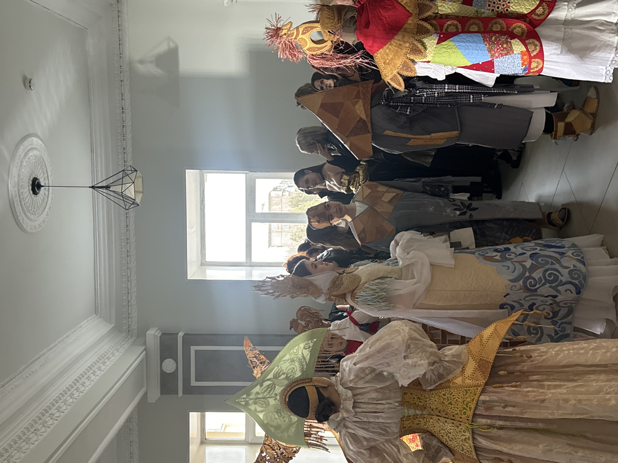 A group of people are standing indoors, possibly in a room with high ceilings and decorative molding, which may be located in an unknown city. They are wearing elaborate costumes with pointed headdresses and patchwork-like garments. A modern chandelier hangs from the ceiling. Natural light is coming through a window in the background.