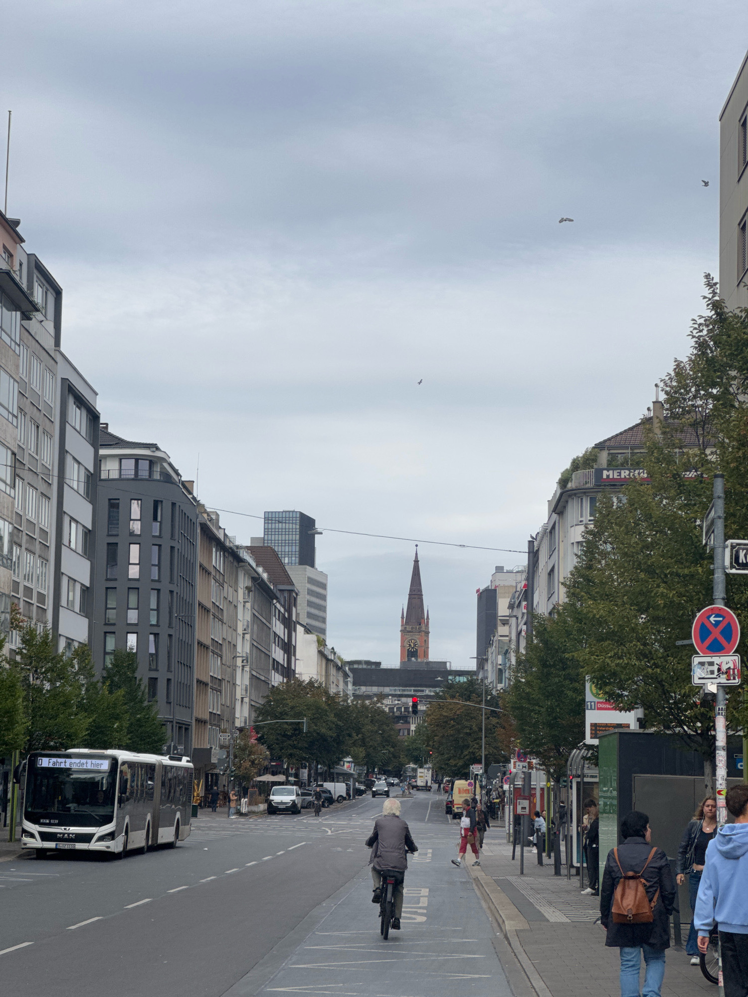 This is a street-level, medium-distance, eye-level, daytime, outdoor, vertical shot of a wide city street in Dusseldorf, Germany. The street is lined with multi-story buildings on both sides. On the left, there are modern apartment or office buildings, one with a light gray facade and numerous windows, and another with a dark gray facade. On the right, the buildings are a mix of architectural styles, including one with a sign that says "MERKUR" and a rooftop terrace with greenery. A tall church steeple with a clock is visible in the distance, centrally located between the buildings. The sky is overcast and cloudy.
Several people are present. A person is riding a bicycle in the middle of the street. Another person in a gray jacket and pants is walking on the sidewalk. A group of people are standing at a bus stop on the right side of the street. A bus with the destination "Fahrt endet hier" (Journey ends here) is driving on the left side of the street. Several other vehicles, including cars and a white van, are also on the road. Traffic lights are visible. Traffic signs, including a red circular sign with a blue arrow pointing left and a red diagonal bar through it, and a rectangular sign with text and an image, are present on the right side. Trees with green foliage are planted along the sidewalk on the right. The street is paved with asphalt, and there are white lane markings and pedestrian crossings.
