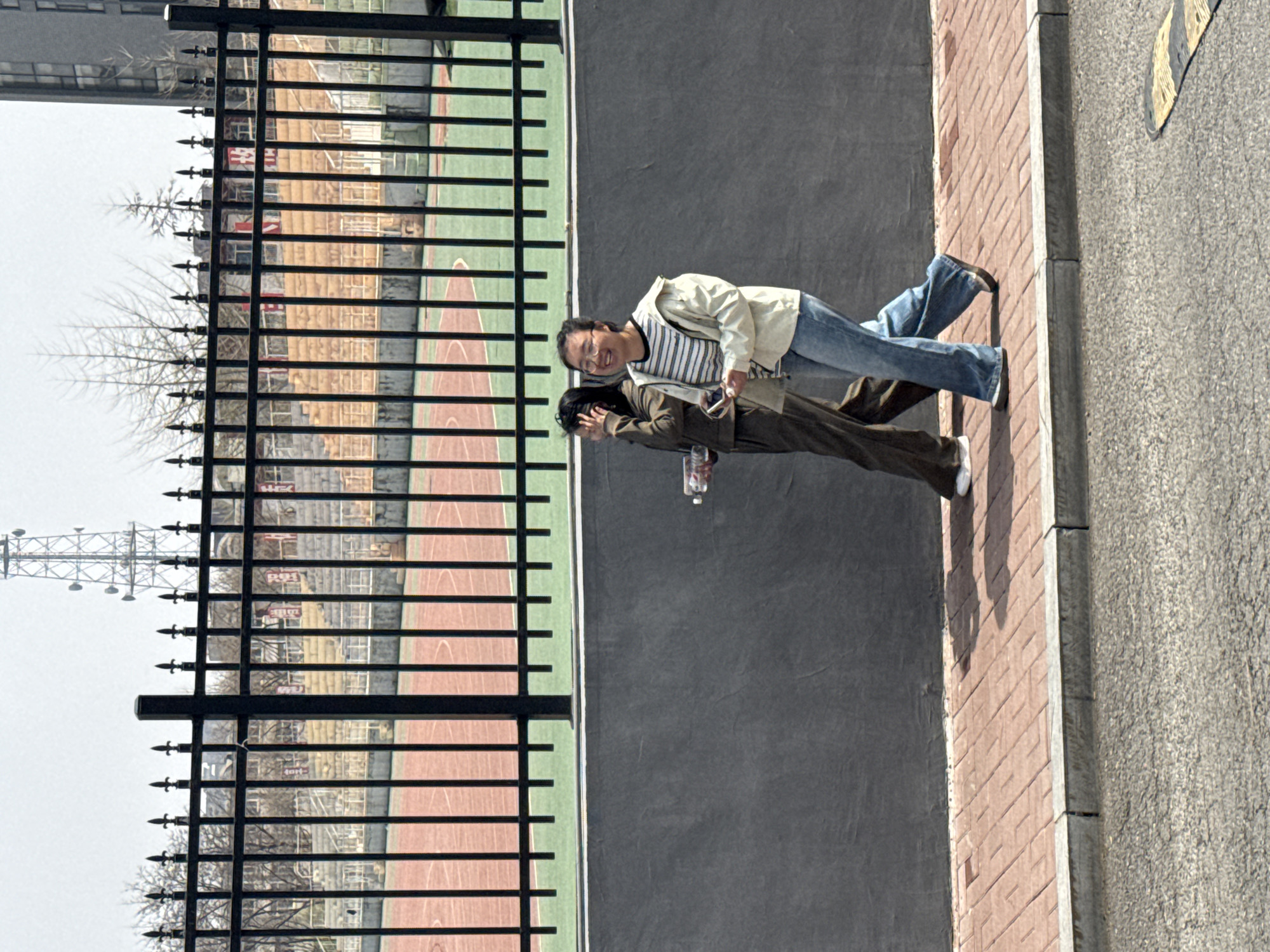 This image, taken in Zhongtun, China, captures an outdoor scene on a clear day. Two people are standing on a brick-paved sidewalk, positioned to the right of a prominent black metal fence that features spear-top finials.
The person on the left, closer to the viewer, wears a light-colored jacket over a striped shirt and blue jeans. They are smiling and looking towards the camera, holding a smartphone in their hand. The second individual, standing slightly behind and to the right, is dressed in a brown jacket and matching pants. Their head is partially obscured or angled downwards, and they are holding a plastic bottle.
The black fence acts as a barrier, separating the sidewalk from a sports court that is visible behind it, characterized by reddish-brown and green sections. In the background, beyond the court, several buildings can be seen. Some of these buildings display faint red characters, likely Chinese, which are unreadable due to distance and resolution. Bare trees are also present in the background, suggesting a season without full foliage. A tall communication tower is visible in the distant background against the bright sky.
To the right of the brick sidewalk, an asphalt road features yellow and black striped markings, indicating a traffic area. The bright, even lighting across the scene suggests it is daytime.