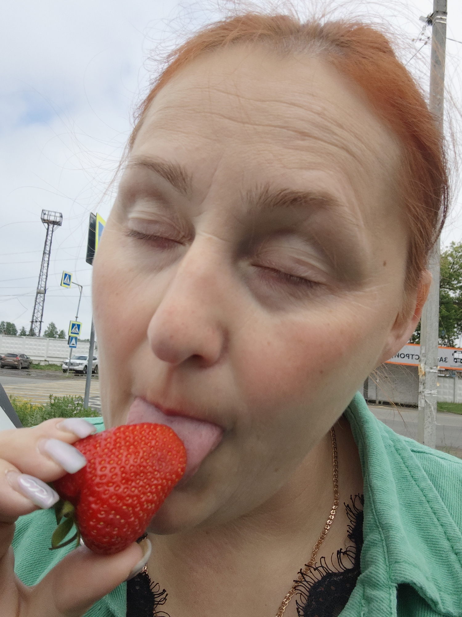 A close-up, slightly low-angle photograph captures a person, appearing female with reddish-auburn hair, licking a red strawberry with closed eyes. This suggests a moment of intense enjoyment or sensory focus. Visible wrinkles are present on the forehead and around the eyes, and the cheeks show a rosy flush. The person wears a green jacket or shirt over a black lace top, accented by a delicate gold chain necklace. Their hand, featuring light-colored, possibly iridescent, manicured nails, delicately holds the ripe strawberry.
The background indicates an outdoor, daytime setting under a cloudy sky, likely in an urban or roadside area. To the left, a tall metal lattice tower, resembling an electricity pylon or communication tower, dominates the distant skyline. Below it, a road is visible, flanked by a low concrete barrier or fence, with several cars in the distance. Two yellow pedestrian crossing signs are also present. To the right, a utility pole stands next to another section of concrete fence. Partially visible behind the pole is a white sign with black Cyrillic text, reading "Я ОСТО..." and "...АЕ", which appears to be part of a bus stop sign or similar public information board, consistent with a location in Chaikovsky, Russia.