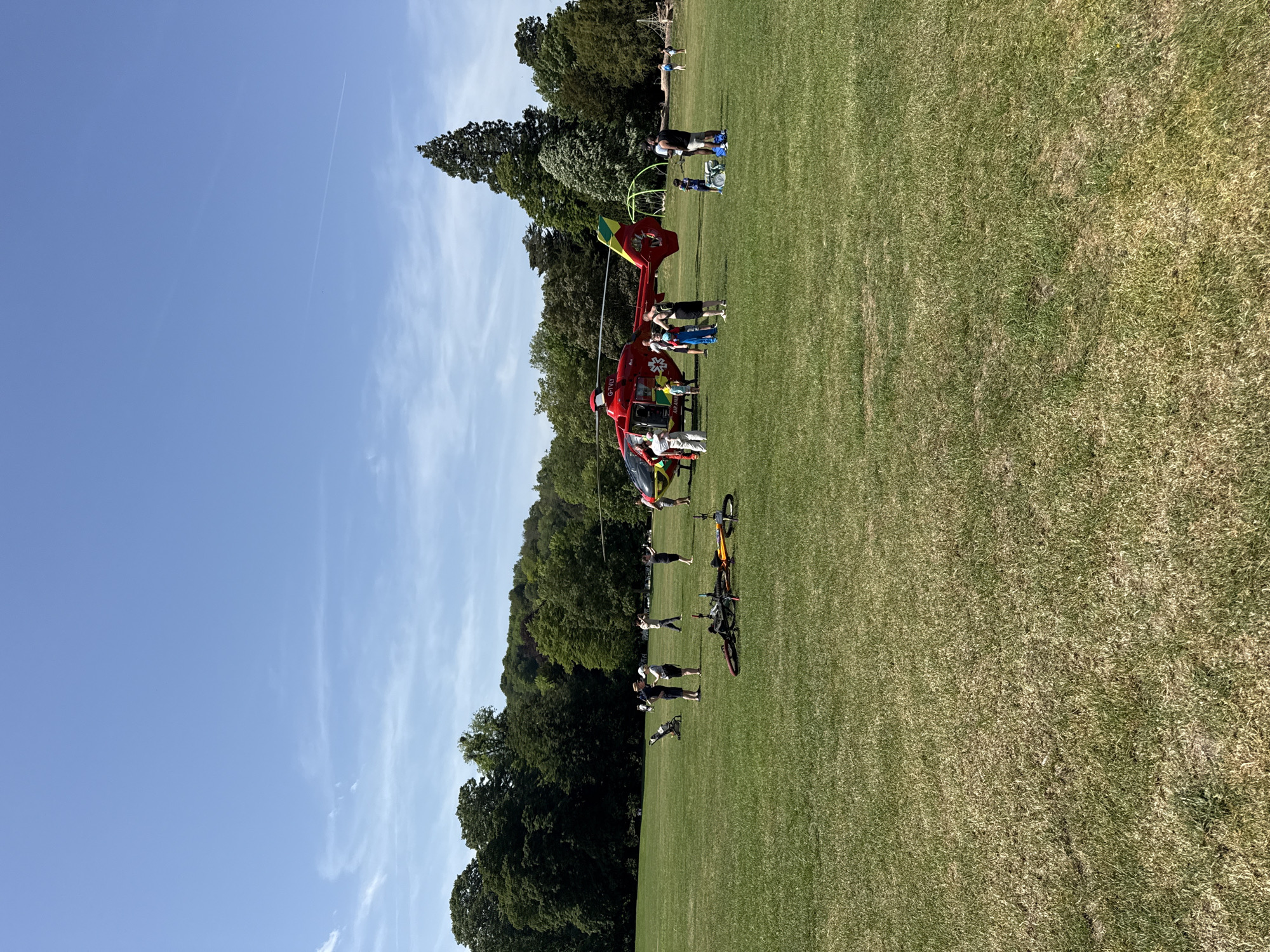 A red and yellow helicopter with the marking "G-TILY" is grounded on a grassy field in High Wycombe, United Kingdom. Multiple individuals are standing around and near the helicopter. In the foreground, there are two bicycles laying on the grass. The background consists of green trees and a blue sky with wispy clouds. Additional individuals can be seen at the far end of the field.