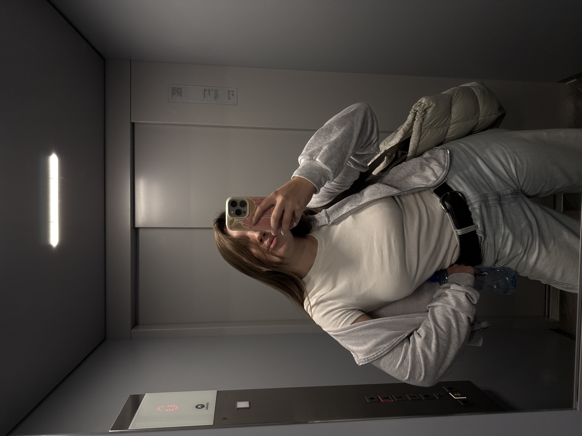 A young woman is taking a selfie in an elevator. She is wearing a white t-shirt, a grey zip-up hoodie, and light-wash jeans with a black belt. She is holding a water bottle in her left hand and her phone in her right. The elevator has grey walls, a vertical rectangular light fixture, and a control panel with buttons labeled "0", "1", "2", and "3". The current floor is highlighted in red. It is unlikely, based on the image, that this scene can be definitively placed within the city of Vozdovac Urban Municipality, Serbia.