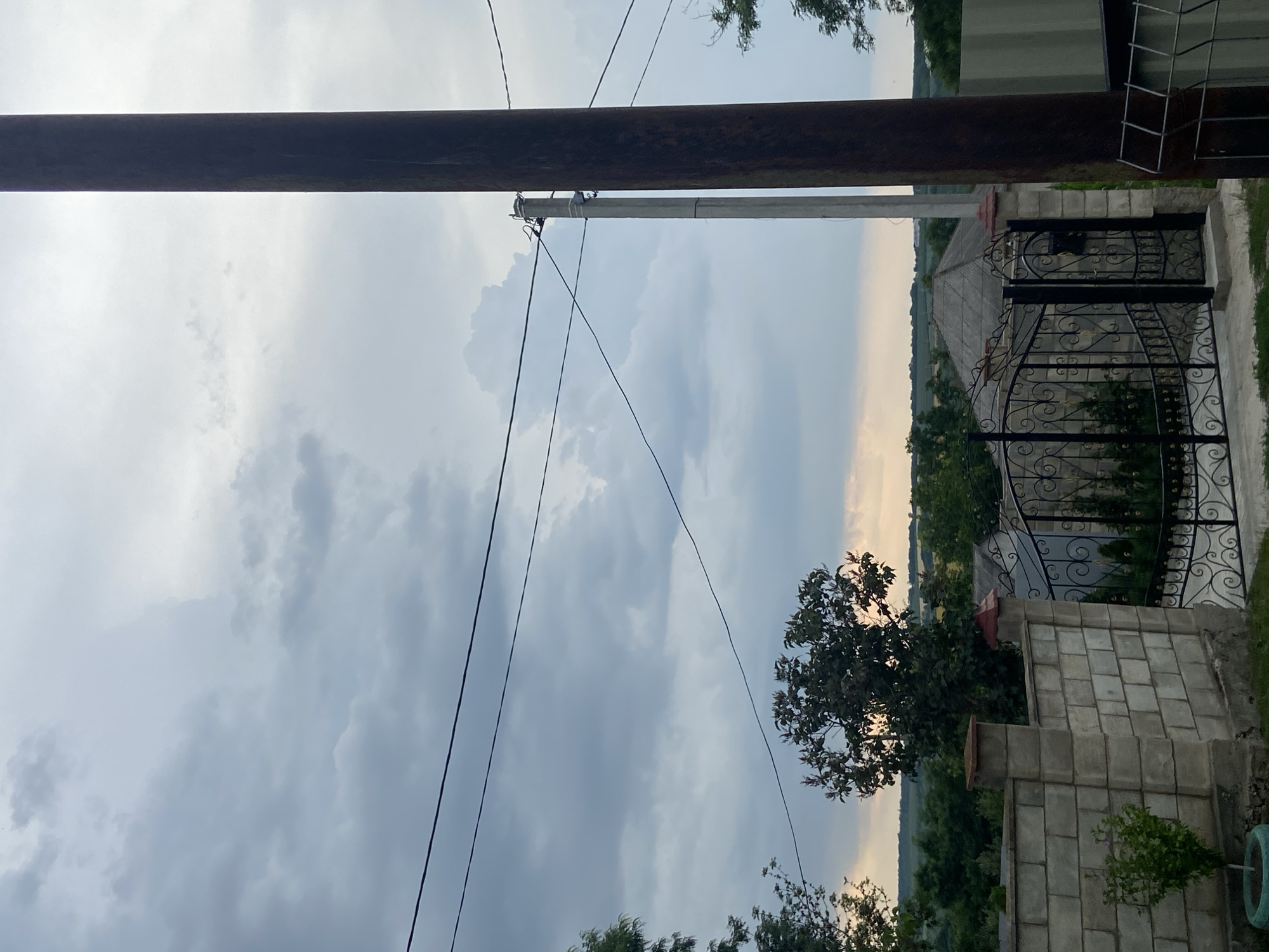 The image shows an outdoor scene with a cloudy sky. In the foreground, there is a tall, dark wooden utility pole on the right and a lighter concrete utility pole to its left, with power lines strung between them. To the left of the poles is a stone block wall with decorative caps on its pillars. Beyond the wall, a black wrought iron gate is visible, leading to a property with a house or building behind it, featuring a grey, pitched roof. The background consists of lush green trees and a rolling landscape under a dramatic, overcast sky that hints at the possibility of rain. The setting appears to be a rural or semi-rural area.