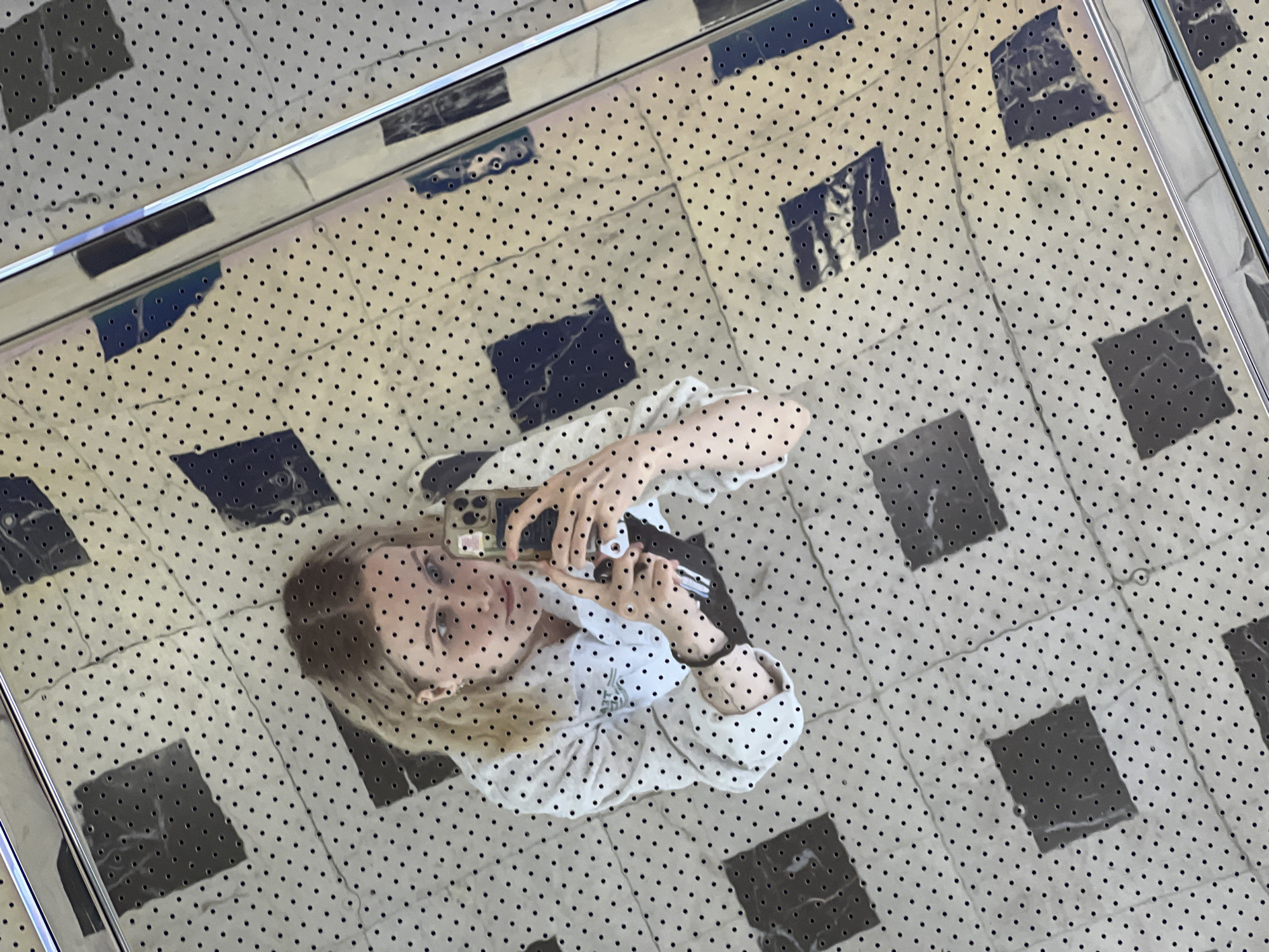 A person is taking a selfie in a mirrored room. The person is a woman with blonde hair, wearing a white patterned shirt. She is holding a smartphone in her right hand and appears to be pointing it at herself. The background is a tiled floor with a pattern of light and dark squares and a grid of small black dots, reflected in a mirror. The scene is indoors and appears to be in a public space due to the reflective nature of the environment. This image was taken in the city of Khimki, Russia.