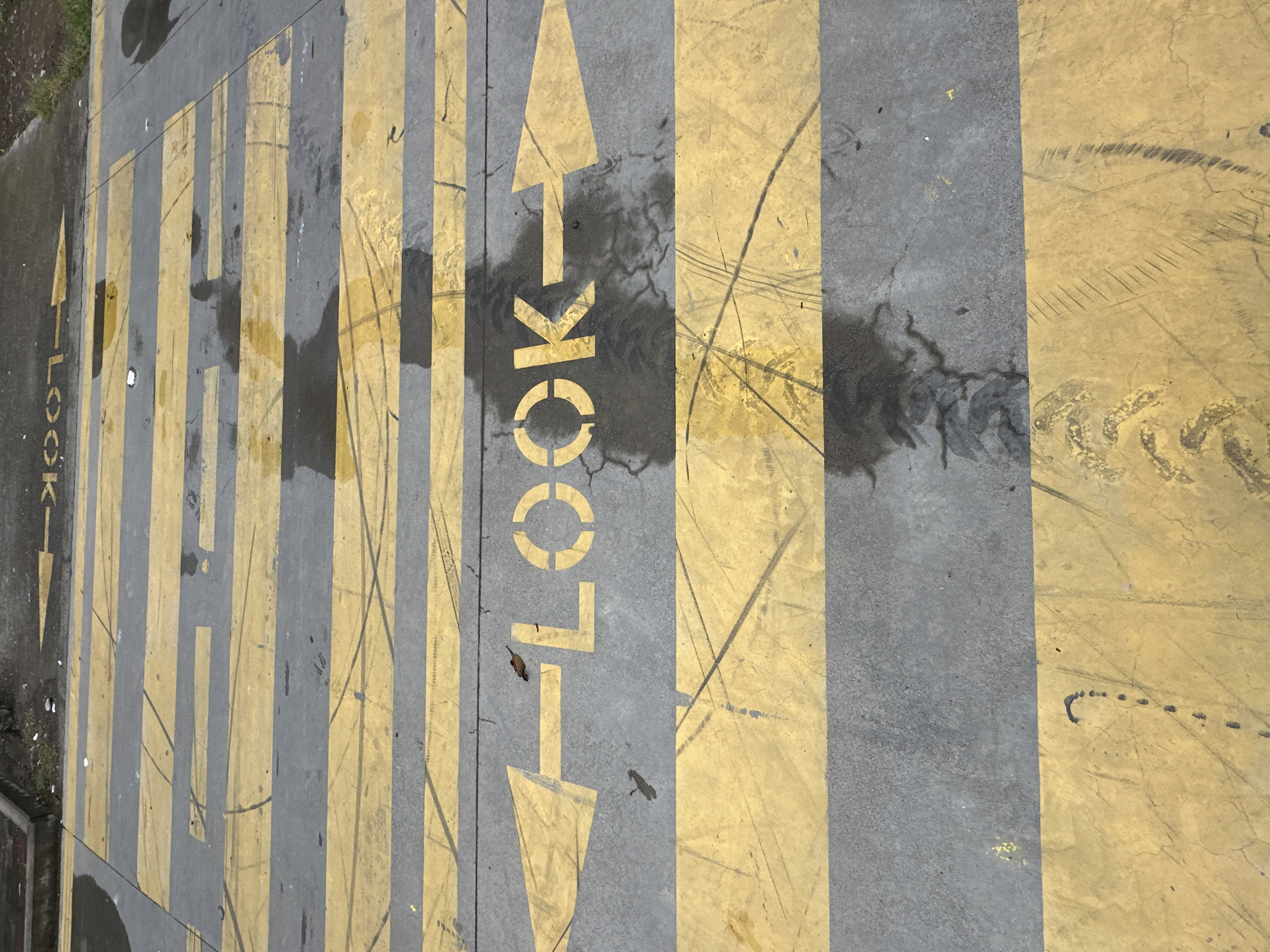 The image shows a section of a grey concrete sidewalk in Batumi, Georgia, featuring yellow painted markings. These markings consist of parallel stripes alternating with grey concrete. Vertical markings read "LOOK" with arrows pointing both upward and downward. There are dark, seemingly wet spots on the surface, as well as tire tracks and other signs of wear and tear.