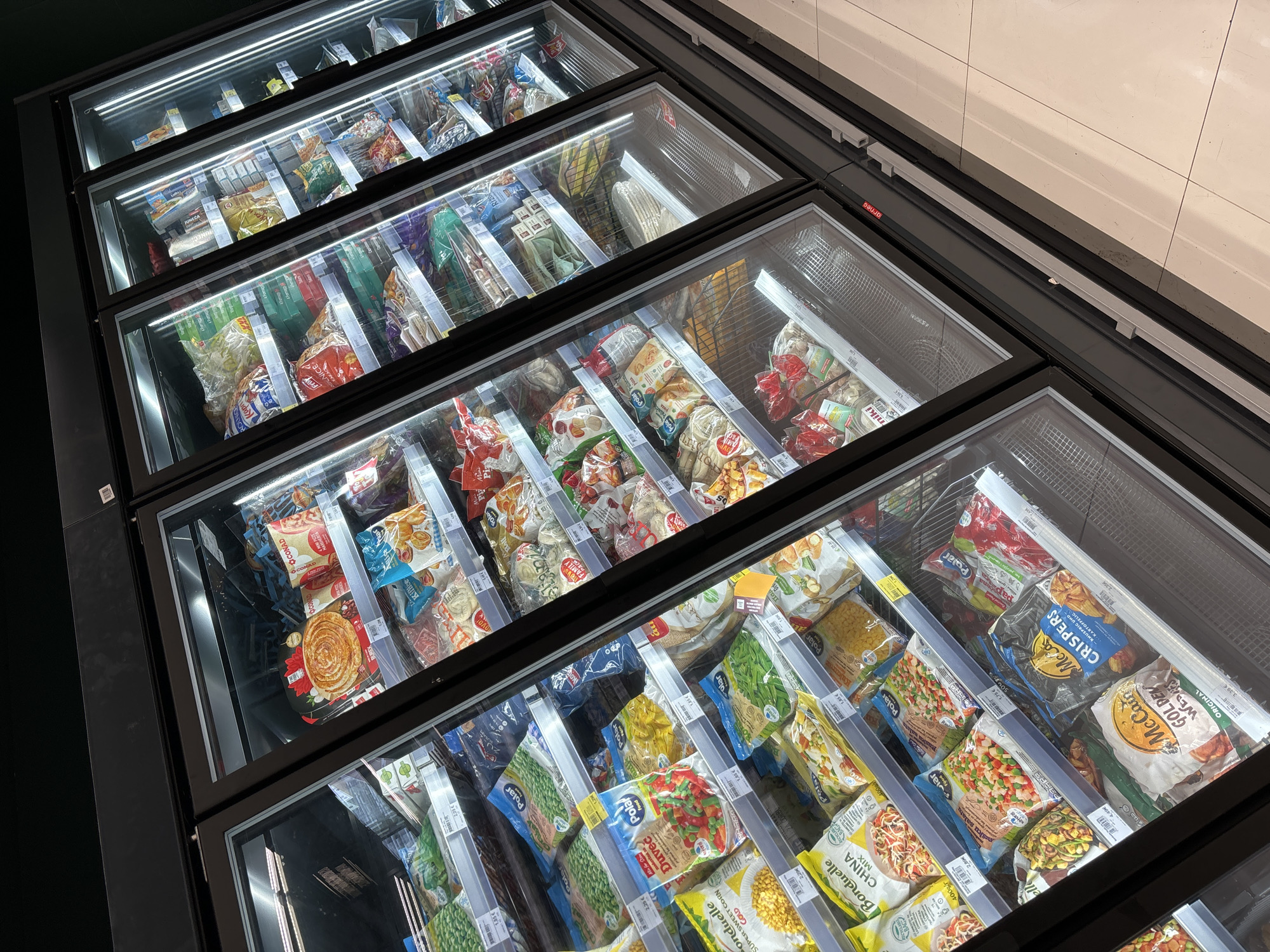 The image shows a well-lit aisle of a supermarket in Budva, Montenegro, with a focus on a large, multi-door refrigerated display case filled with frozen food products. The display case is divided into multiple vertical sections, each showcasing a variety of packaged frozen items such as vegetables, pizzas, and other convenience foods. The products are neatly arranged on shelves, with price tags visible on most of them. The lighting from within the display case creates a bright and appealing presentation of the merchandise. The floor in front of the display case is tiled and appears clean. There are no people visible in the immediate vicinity of the display case. The overall environment suggests a typical retail setting in a grocery store.
