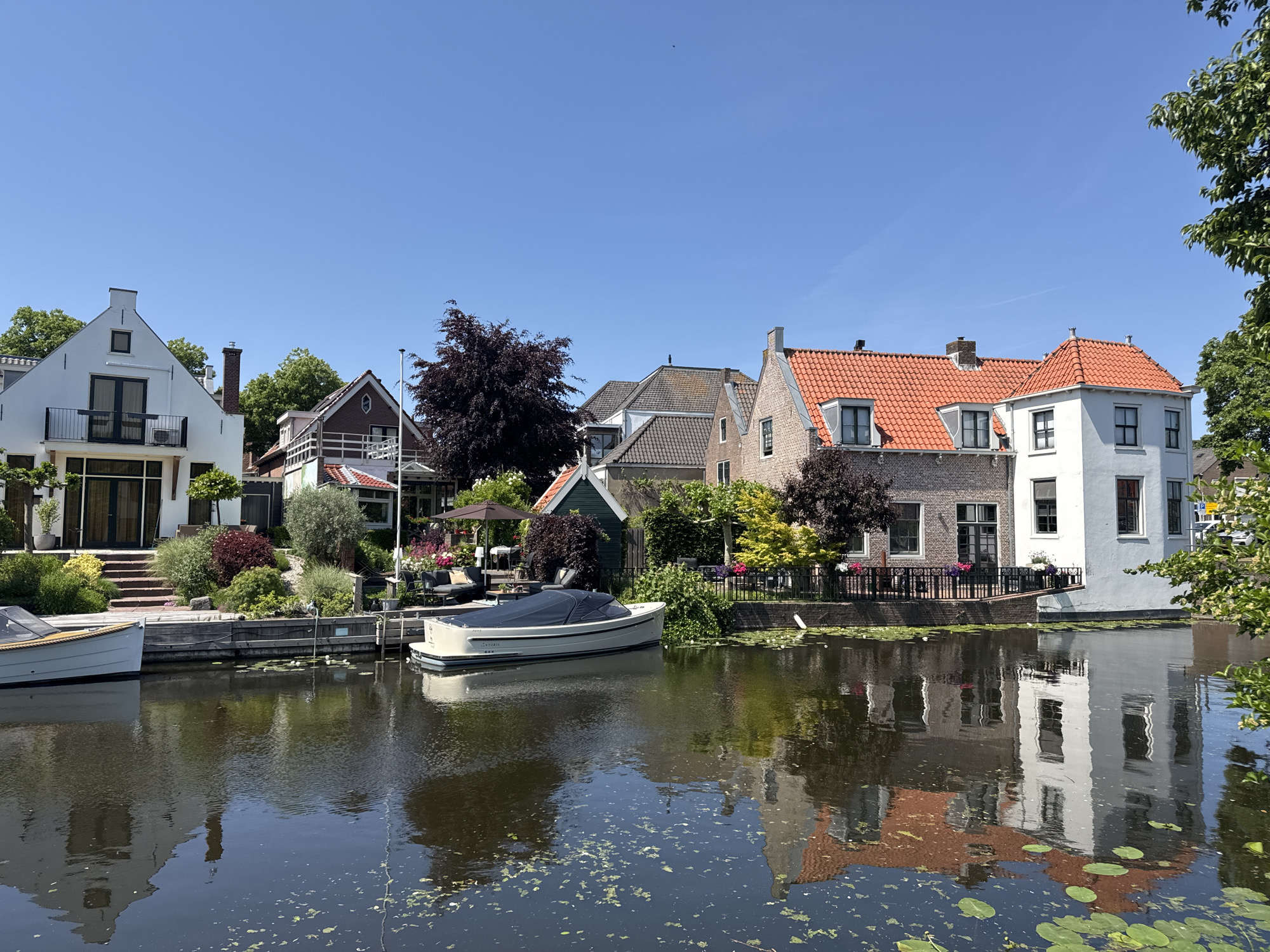 The image shows a canal in Katwijk, Netherlands, lined with houses and boats. The houses vary in style, with some being white with gabled roofs and others having red-tiled roofs and brick facades. A boat is docked along the canal bank, covered with a dark blue tarp. The canal is filled with water and some aquatic plants or algae. Reflections of the buildings are visible in the water. The sky is clear and blue. The scene depicts a quiet, residential area.