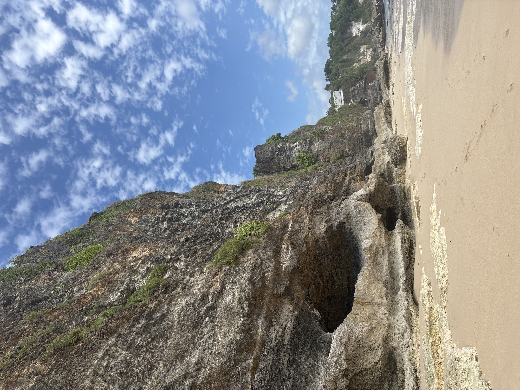 The image depicts a sandy beach in Kuta Selatan, Indonesia, with a dramatic, sheer cliff face dominating the left side of the frame. The cliff is composed of layered rock, with visible striations and textures, and is partially covered with green vegetation, including small trees and shrubs clinging to its surface. Several natural openings and eroded sections are present in the cliff base, forming small caves and overhangs.
The beach itself is composed of light brown sand, with gentle waves lapping at the shore on the right side of the image. Footprints are visible in the sand, indicating recent human presence. In the distance, along the crest of the cliff, a building with white signage is visible, suggesting human settlement or a resort in the area. The sky above is a vibrant blue, scattered with fluffy white clouds, indicative of a clear and sunny day. The overall scene captures the natural beauty of a tropical coastline.