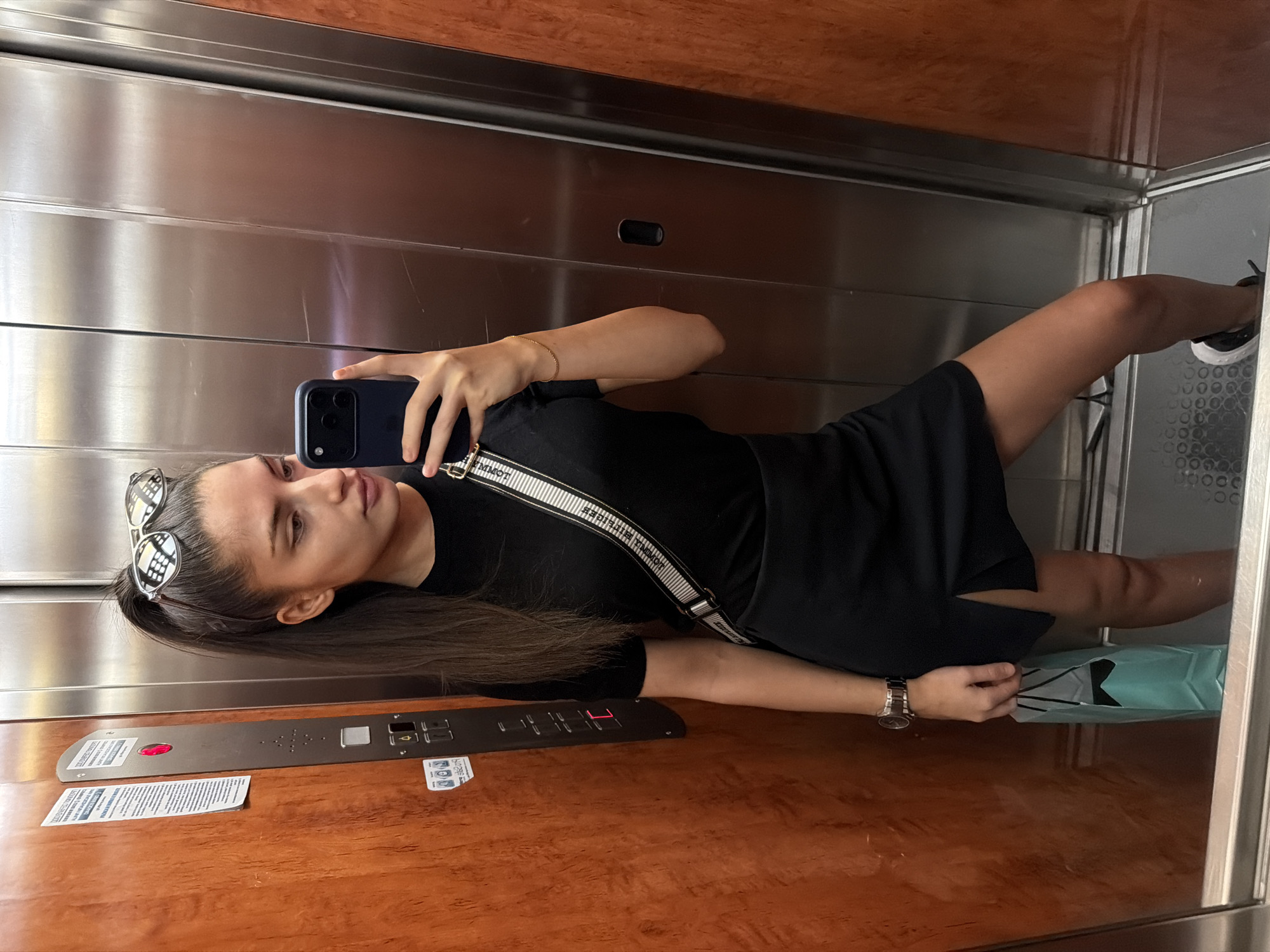 The image shows a young woman in an elevator, taking a selfie. She is wearing a black t-shirt and a black skirt with a thigh-high slit. A white strap with black lettering crosses her chest, and she is holding a dark blue smartphone in her right hand. A silver watch is visible on her left wrist, and she is carrying a light blue shopping bag in the same hand. She has her hair pulled back in a ponytail and is wearing sunglasses on top of her head. The elevator has wooden paneling on the left wall and a stainless steel door and wall on the right. The elevator control panel on the left wall has several buttons and a red illuminated button. The floor of the elevator appears to be metallic with a textured pattern. The scene is likely a public elevator within a building in the city of Plovdiv, Bulgaria, though the exact location within the city is not specified.