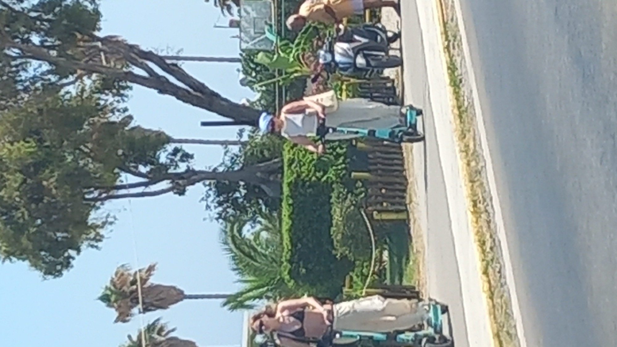 The image captures a sunny outdoor scene with people and vehicles on a paved road, likely in Heraklion, Greece, given the presence of palm trees and Mediterranean vegetation. In the foreground, a light-colored road is visible with a yellow and white border. Further back, two individuals are riding electric scooters. The person on the left, wearing a black bikini top and light-colored pants, is on a teal scooter. To their right, another person, dressed in a white top and light pants and wearing a blue headscarf, is on a similar teal scooter. Behind them, to the right, a dark-colored motorcycle is parked, and a man in a yellow shirt and shorts stands nearby. A decorative wooden fence lines the edge of the road, beyond which a well-maintained hedge and various trees, including palm trees, are visible under a clear blue sky.