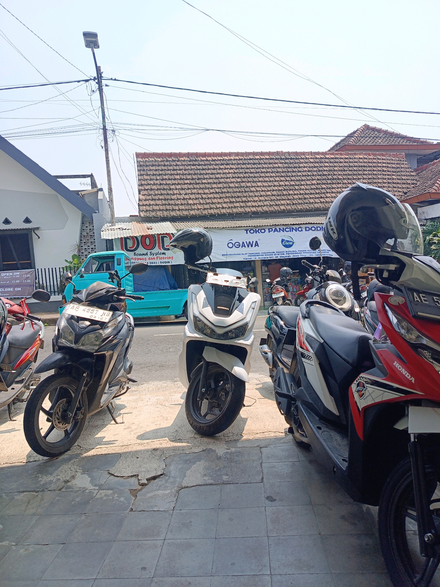 The image captures a street scene in front of a shop in Kebonagung, Indonesia. A light blue pickup truck is parked on the street, partially obscured by several parked motorcycles. The motorcycles are lined up along the sidewalk, with their fronts facing the viewer. One motorcycle, a white Honda, is prominently in the foreground on the right, its red and white bodywork visible. Next to it is a black motorcycle, also a Honda, with its license plate reading "AB 4557 MB". Another white motorcycle is visible to the left of the black one. A black helmet rests on the handlebars of the red and white Honda. The shop behind the motorcycles has a tiled roof and a banner that reads "TOKO PANCING DODI" and "OGAWA". Power lines crisscross the sky above the scene, and a street light is visible on a pole. The sidewalk appears to be paved with concrete tiles, some of which are cracked. The environment suggests a commercial area with typical street infrastructure.