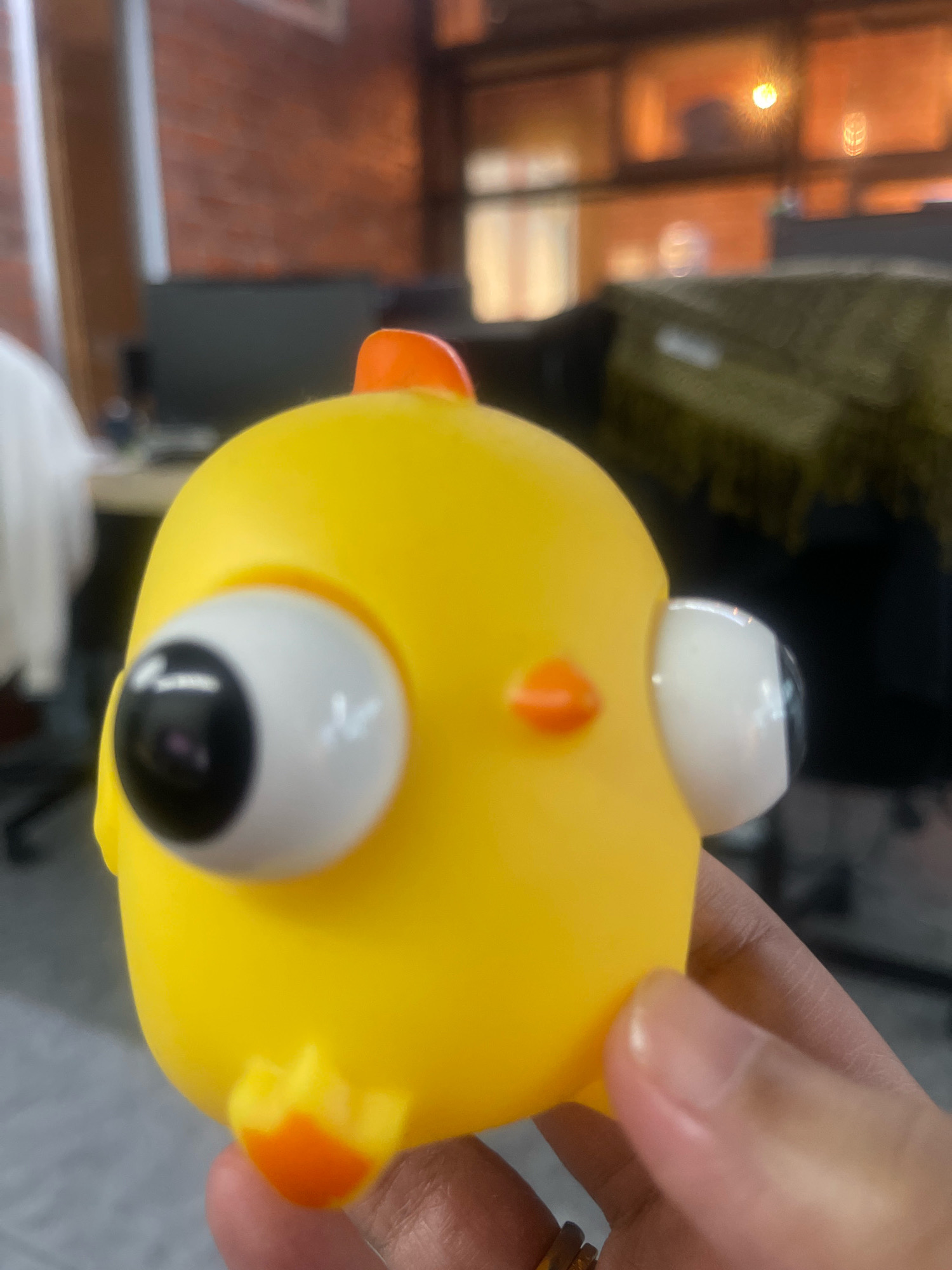 A close-up, slightly tilted, eye-level shot focuses on a bright yellow, squeezy toy chick held by a person's hand. The chick is the primary subject, with its large, white, bulgy eyes featuring black pupils and prominent white reflections. It has a small orange beak and a small orange crest on its head. Another orange protrusion, possibly a leg or part of its body, is visible near the bottom. The background is blurred but suggests an indoor setting, possibly an office environment, with hints of brick walls, shelving units, and a patterned green fabric. The lighting is warm, with a visible light source reflecting in the chick's eyes and the background. The image quality is slightly soft. The location is Subang Jaya City Council, Malaysia.