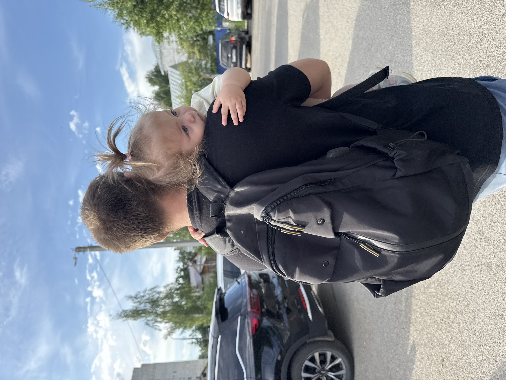 A man is holding a young child who is leaning on his shoulder. The man has a backpack on. The scene appears to be outdoors, possibly a parking lot, in the city of Chaikovsky, Russia. Cars are visible in the background.