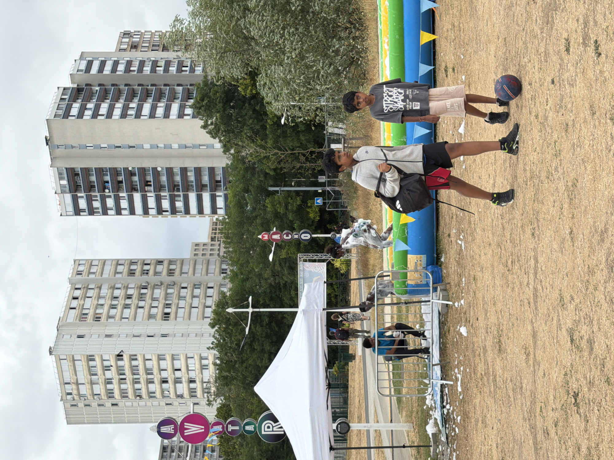 The image depicts an outdoor scene in the city of Bobigny, France, likely within a residential area given the presence of tall apartment buildings in the background. In the foreground, two young boys are walking on dry, sparse grass. The boy on the left, wearing a grey hoodie, black shorts, and black sneakers with green accents, carries a black backpack over his shoulder. The boy on the right, wearing a grey t-shirt with "STREET SKATER" printed on it and light brown shorts, stands near a soccer ball. Behind the boys, a large inflatable structure with yellow and blue sections and colorful triangular flags is visible, suggesting a recreational event or park activity. A white pop-up tent with metal poles is set up to the left, partially obscuring a metal barrier. A person in a blue t-shirt is seated behind the barrier. In the mid-ground, more people are visible, some walking and others near what appears to be an inflatable slide or obstacle. Several tall, rectangular apartment buildings dominate the background, with visible windows and balconies. Circular signs with letters are mounted on poles in the distance, one displaying "A V A T A R" and another "M A C H O". The sky is overcast.
