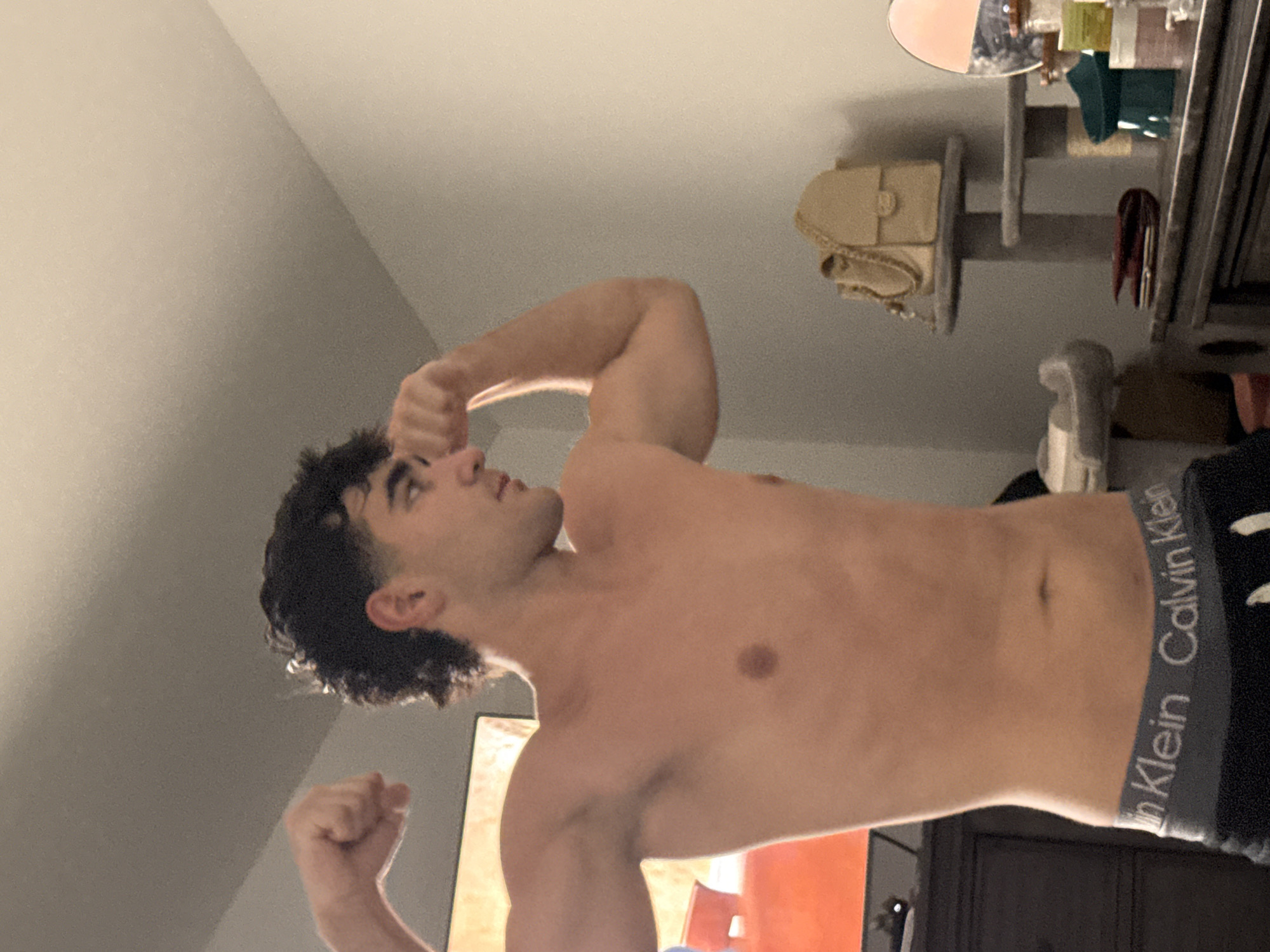 A young, shirtless man with dark, curly hair is flexing his biceps in a dimly lit room. He is wearing Calvin Klein underwear. The background shows a dresser, a television, and a shelf with a purse, a mirror, and various containers. The scene appears to be indoors, likely a bedroom, in Bellmore, United States of America.