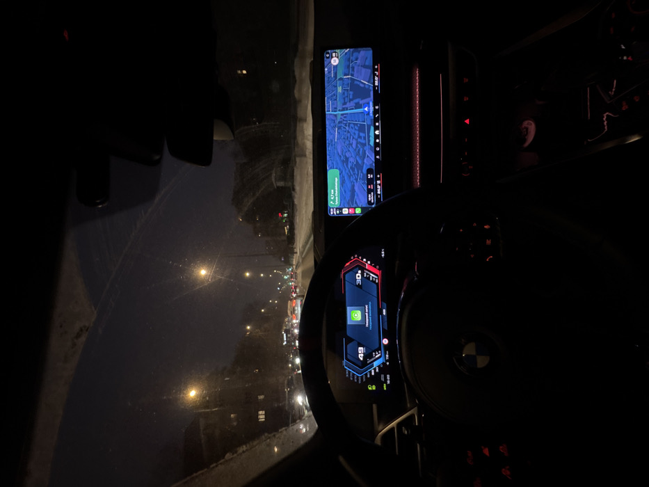 The image depicts the interior of a vehicle, likely a BMW, viewed from the driver's perspective at night during winter conditions in Warsaw, Poland. Snow and ice residue are visible on the windshield. The digital instrument cluster displays a speed of "49" (likely km/h), a green icon with Polish text "Nieznany znak" (unknown sign), and ambient red lighting on the right side. The infotainment screen to the right shows a 3D navigation map indicating a destination "0.7 km" away, with the visible street name "Puławska" and a branching street appearing to be "Rzymowskiego", consistent with the Ursynów district of Warsaw. Outside the vehicle, a multi-lane city street is visible under a dark sky, illuminated by numerous streetlights. Traffic consists of multiple vehicles with headlights and taillights visible, appearing to be in a queue. Snow covers the ground along the roadside, and bare trees are present on both sides of the street. Low-rise buildings are discernible in the background to the left. No individuals are explicitly visible.