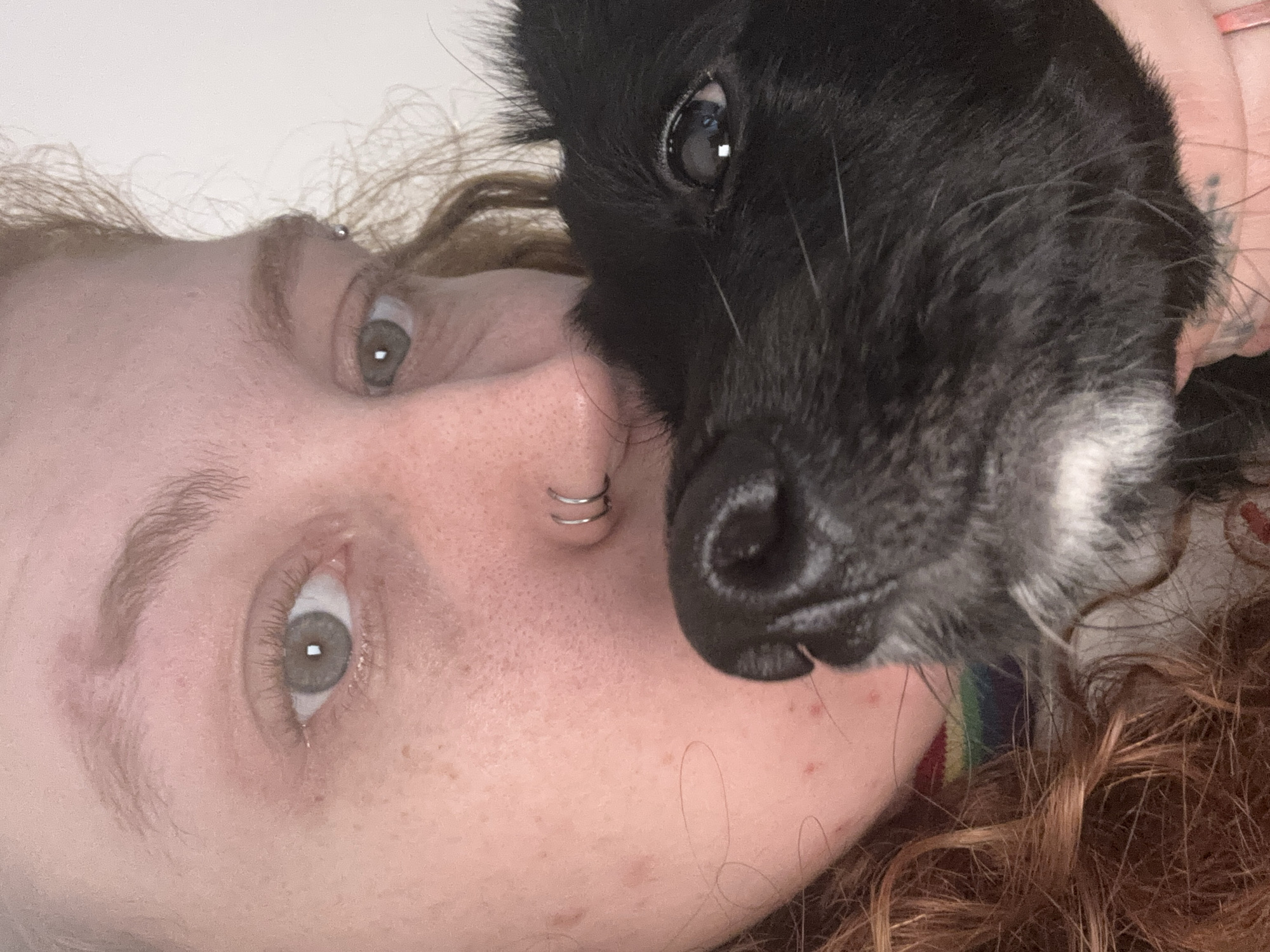 A close-up, slightly rotated image shows a person with freckled skin and curly reddish-brown hair lying down with a black dog. The person has light gray eyes and a double nose ring. The dog, positioned close to the person's face, has a mostly black coat with some white fur around its muzzle. A hint of a rainbow-colored object is visible near the person's shoulder. The background appears to be a plain white surface. The location and the city of Buenos Aires, Argentina cannot be determined from the image.