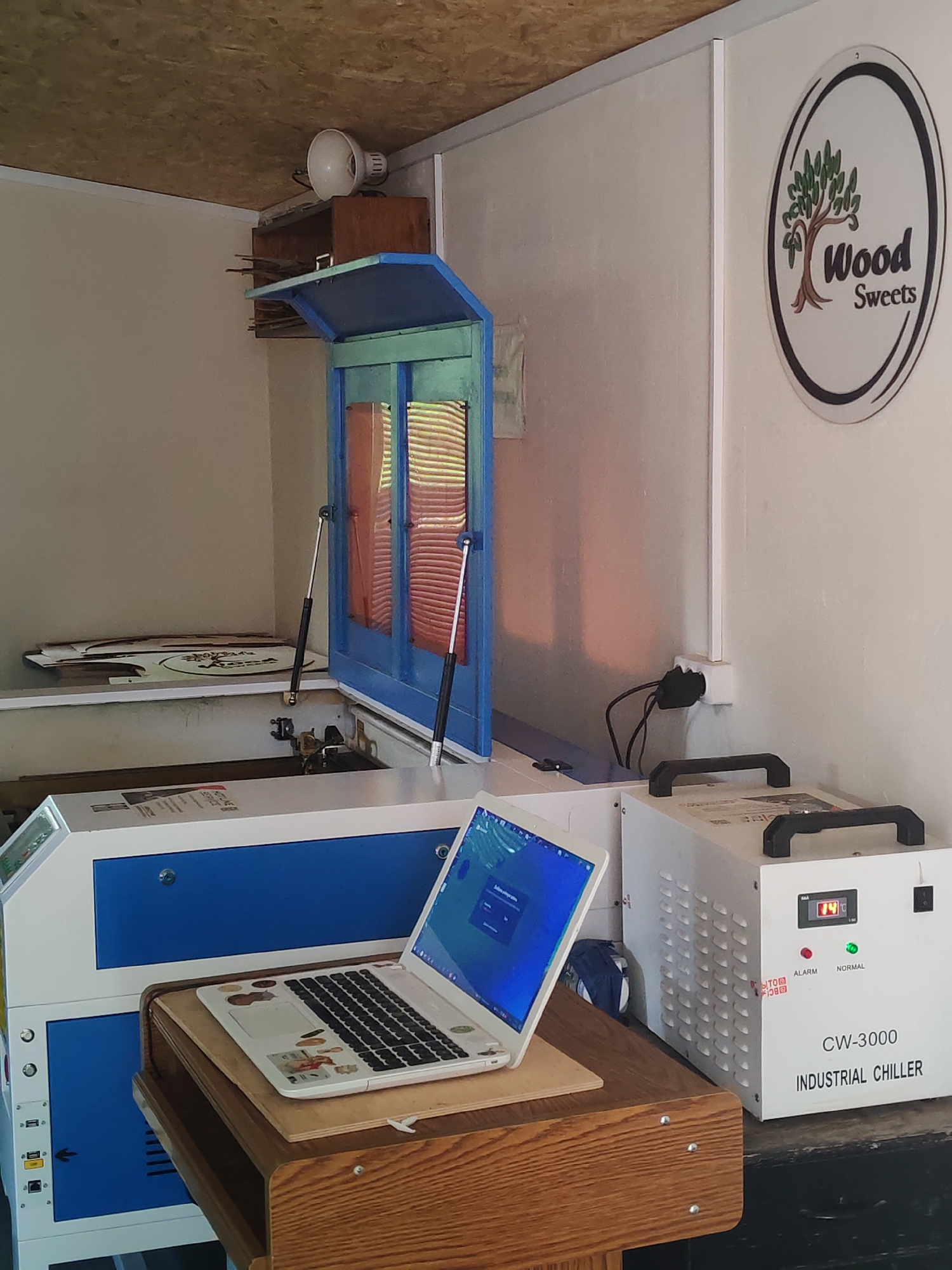 A small room in Unknown, Unknown, houses a laser cutting setup. The focal point is a blue and white laser cutting machine with its lid open. A white laptop rests on a wooden stand in front of the machine. To the right is a white CW-3000 Industrial Chiller. The walls are painted white, and a "Wood Sweets" logo is mounted on the right wall. The lighting is bright, likely artificial. A wooden shelf above the laser cutter holds various items, including a white spotlight. An electrical outlet is visible on the wall near the chiller, and a bag is tucked behind the laptop.