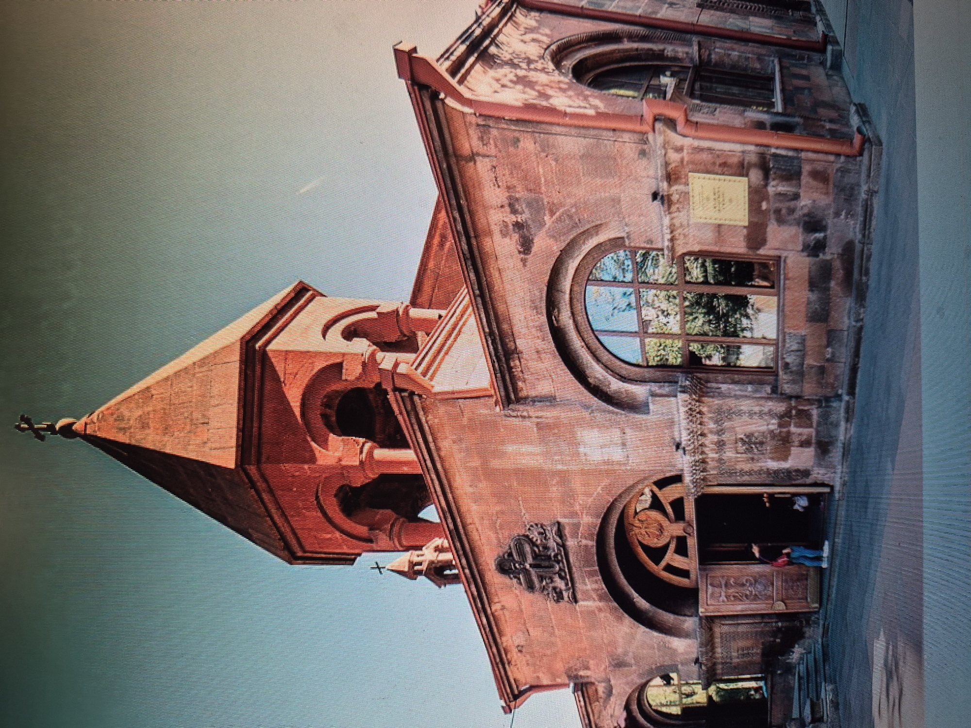 The image depicts the Saint Anna Church in Yerevan, Armenia. The church is constructed of red brick with a prominent conical spire topped with a cross. Arched windows and doorways are visible, and a plaque is mounted on the side. Two people are standing near the entrance, one wearing a red top and blue pants. The building is oriented vertically in the image, with the sky visible behind the spire.
