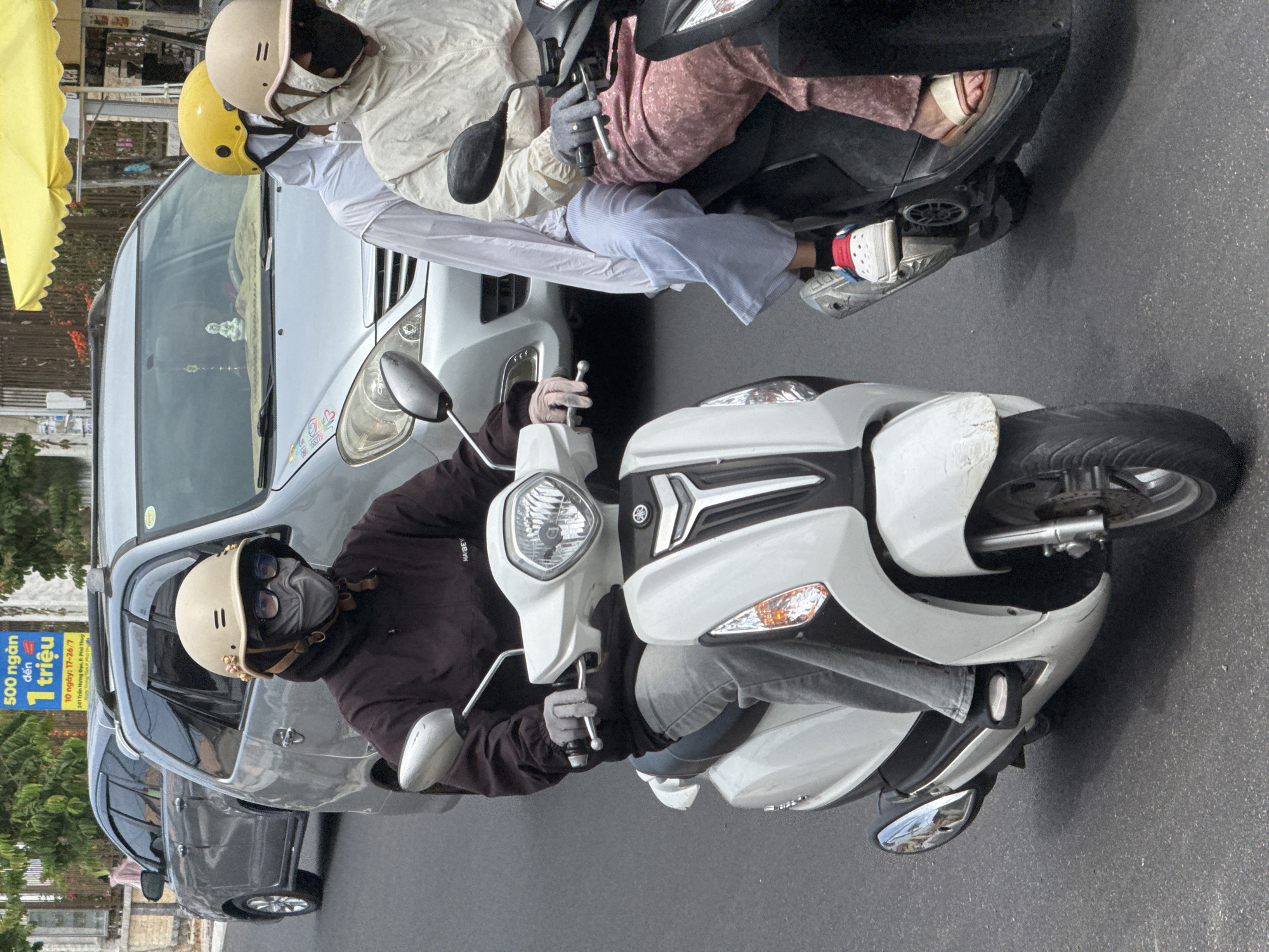 A person wearing a beige helmet, a dark brown hoodie, grey pants, and grey gloves rides a white scooter on a paved road. The person's face is covered with a grey mask and sunglasses. In the background, a silver minivan is parked, and to its right, another person wearing a yellow helmet and a light-colored jacket rides a black scooter. The environment appears to be a street in Phú Thủy, Vietnam, indicated by a sign in Vietnamese in the background that reads "500 ngan den 1 trieu" and "10 ngay: 17-26/7". The sky is overcast.