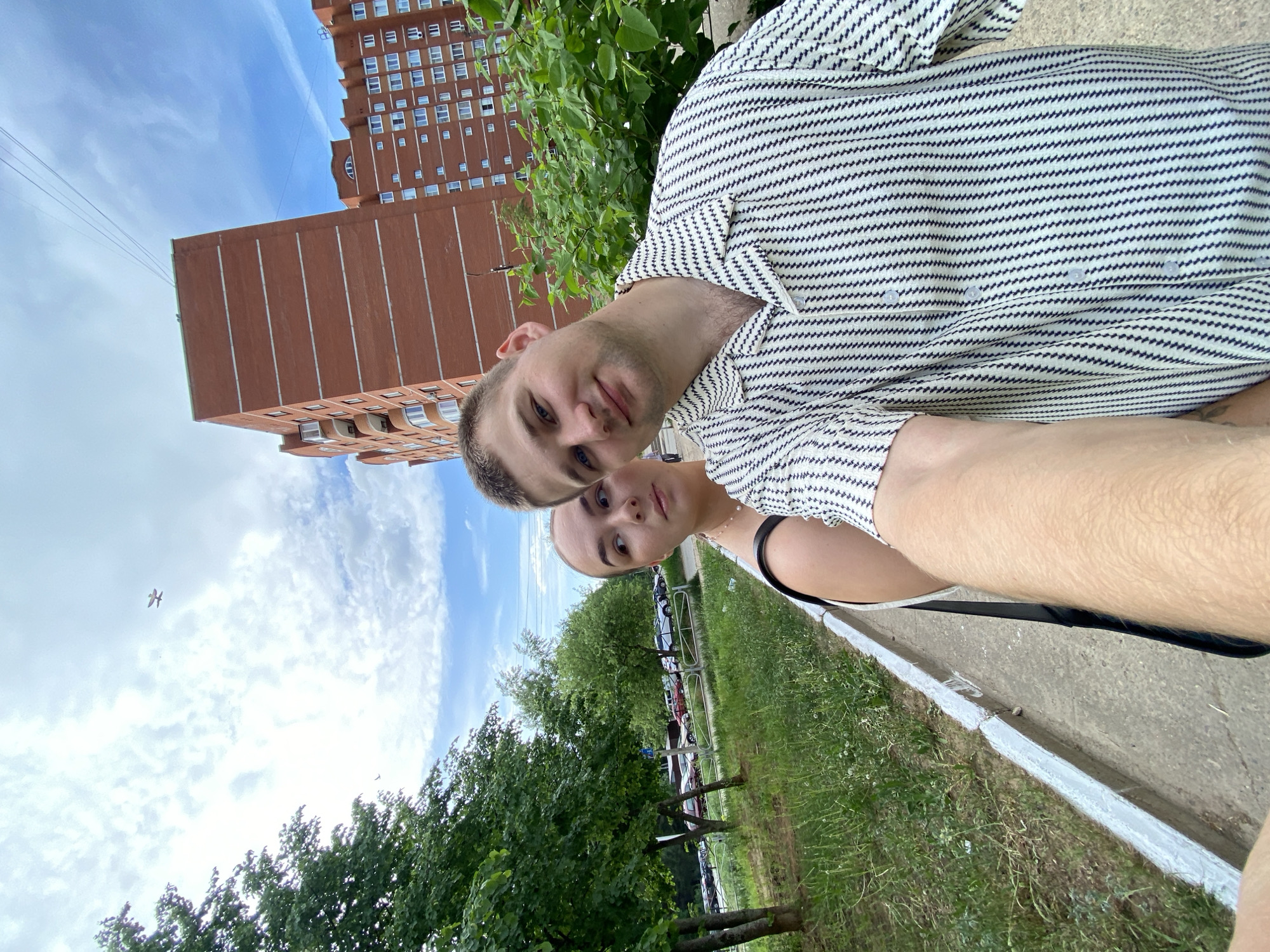 The image shows two people, a man and a woman, standing close together, likely taking a selfie. The man is wearing a striped shirt. A brick building is visible in the background. The location is likely in Chaikovsky, Russia, given the prompt. There are trees and a curb visible, suggesting an urban park or sidewalk setting. A small plane is visible in the sky.