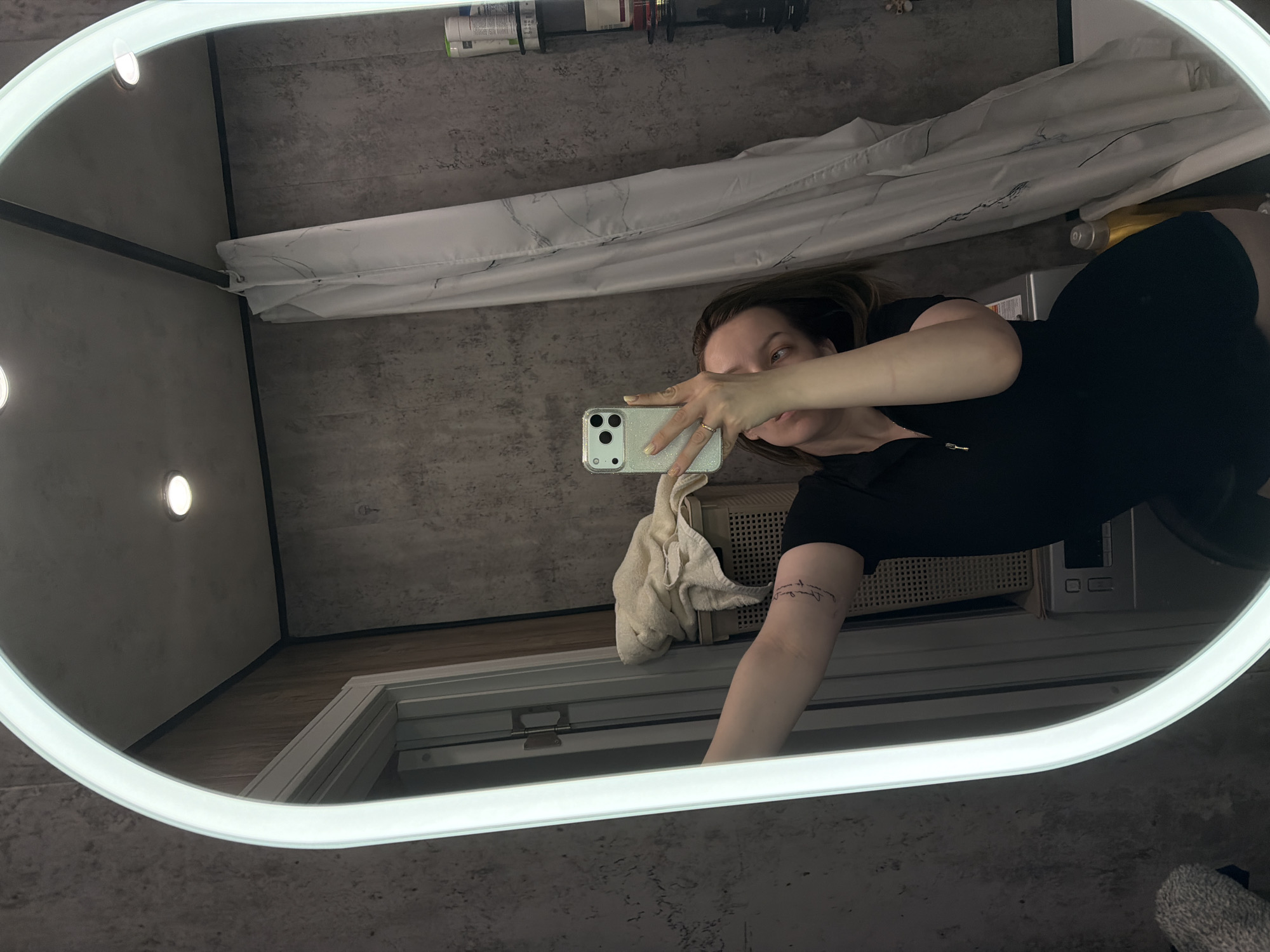 A person is reflected in a lighted oval mirror in what appears to be a bathroom. The person is holding a phone and taking a picture in the mirror. The reflection also shows a shower curtain, toiletries on a shelf, a towel, a floor mat, and what appears to be a vent. The location in Reshetnikovo, Russia, is not verifiable from the image.