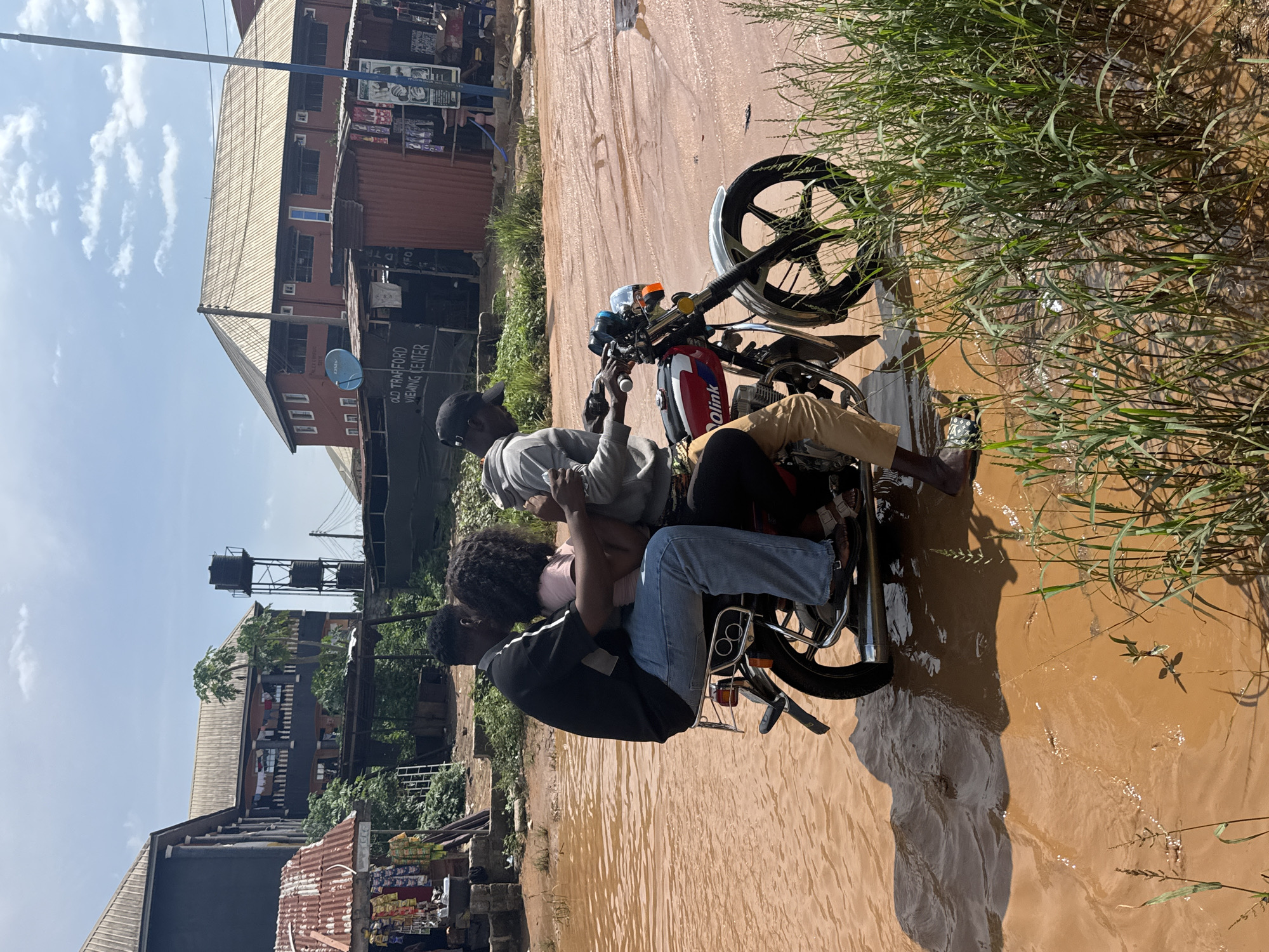 The image depicts a vibrant, challenging scene on a waterlogged, muddy road in Ihiagwa, Nigeria, under bright daylight. Four individuals are situated on and around a red and white motorcycle, which is partially submerged in the extensive brown water covering the ground.
The person at the front, presumably the driver, wears a light grey shirt and a black cap, holding the motorcycle's handlebars. Behind them, a passenger with dark, curly hair is seated, wearing a pink top. A third individual in a black t-shirt and blue jeans sits behind the second passenger, and a fourth person wearing beige pants is positioned further back on the motorcycle. The group appears to be navigating or paused amidst the difficult, water-filled terrain.
To the right of the frame, green grass and weeds grow along the edge of the muddy water. The background features several buildings, indicating an urban or semi-urban environment. One building prominently displays a satellite dish with "DStv" visible. Below it, a sign clearly reads "OLD TRAFFORD VIEWING CENTER." Another building has a corrugated metal roof, and in the distance, multiple water tanks are elevated on a metal stand. The clear blue sky has a few wispy white clouds, and the bright sun casts distinct shadows, emphasizing the intensity of the daylight. The motorcycle's tank is marked with "DalinK."