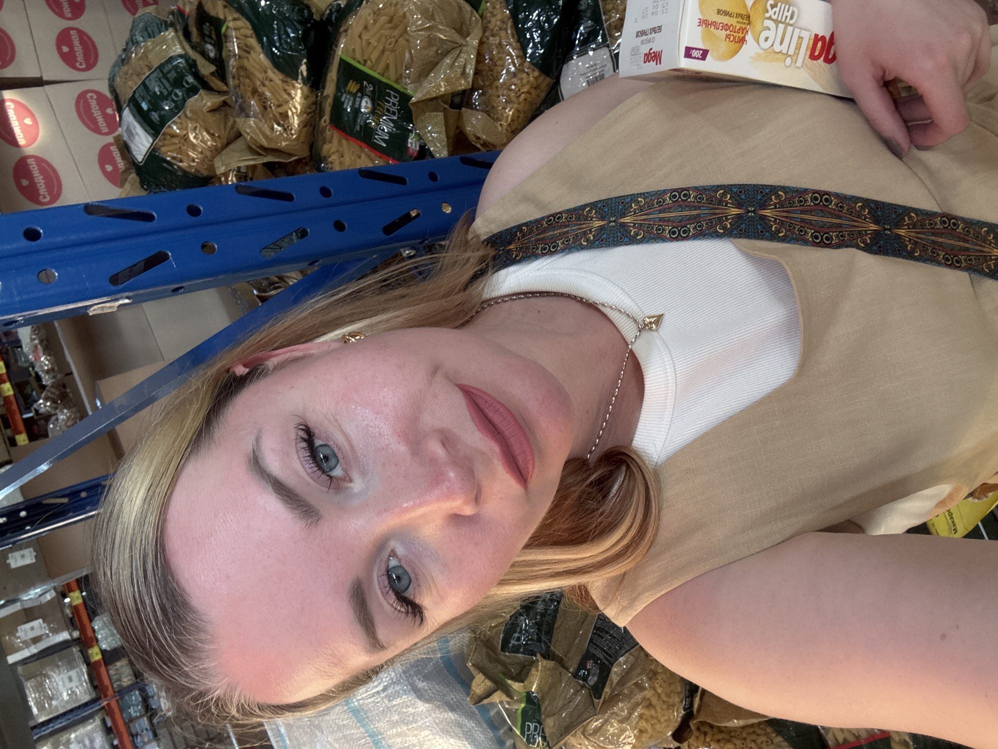 The scene is an indoor retail or warehouse environment, likely a supermarket or large grocery store, located in Панковка, Russia. A fair-skinned woman with blonde hair, styled away from her face, and light blue eyes looks directly at the camera. She wears makeup, including dark eyeliner, extended false eyelashes, and a matte rose-colored lipstick. Her expression is neutral with slightly pursed lips. She is wearing a light-colored (possibly white) ribbed top underneath a tan or beige sleeveless garment, possibly a dress or jumpsuit, featuring a vertical dark blue and orange patterned strap on the right side of her chest. She has a delicate silver-colored chain necklace with a small heart-shaped pendant and small gold-colored earrings.
To her right, partially visible, she holds a white rectangular box of "Mega Line CHIPS," with the flavor indicated as "WHITE MUSHROOM" ("БЕЛЫХ ГРИБОВ") and a weight of "200г." The background features industrial-style blue metal shelving units. On these shelves, numerous large clear plastic bags of dry pasta (appears to be penne or a similar short cut) are stacked. Some pasta packaging displays the word "PREMIUM." Above these, cardboard boxes are visible, several bearing the Cyrillic word "Сладкая" (Sladkaya - meaning "Sweet"), indicating confectionery or sweet products. The overall lighting suggests artificial illumination typical of a retail space.