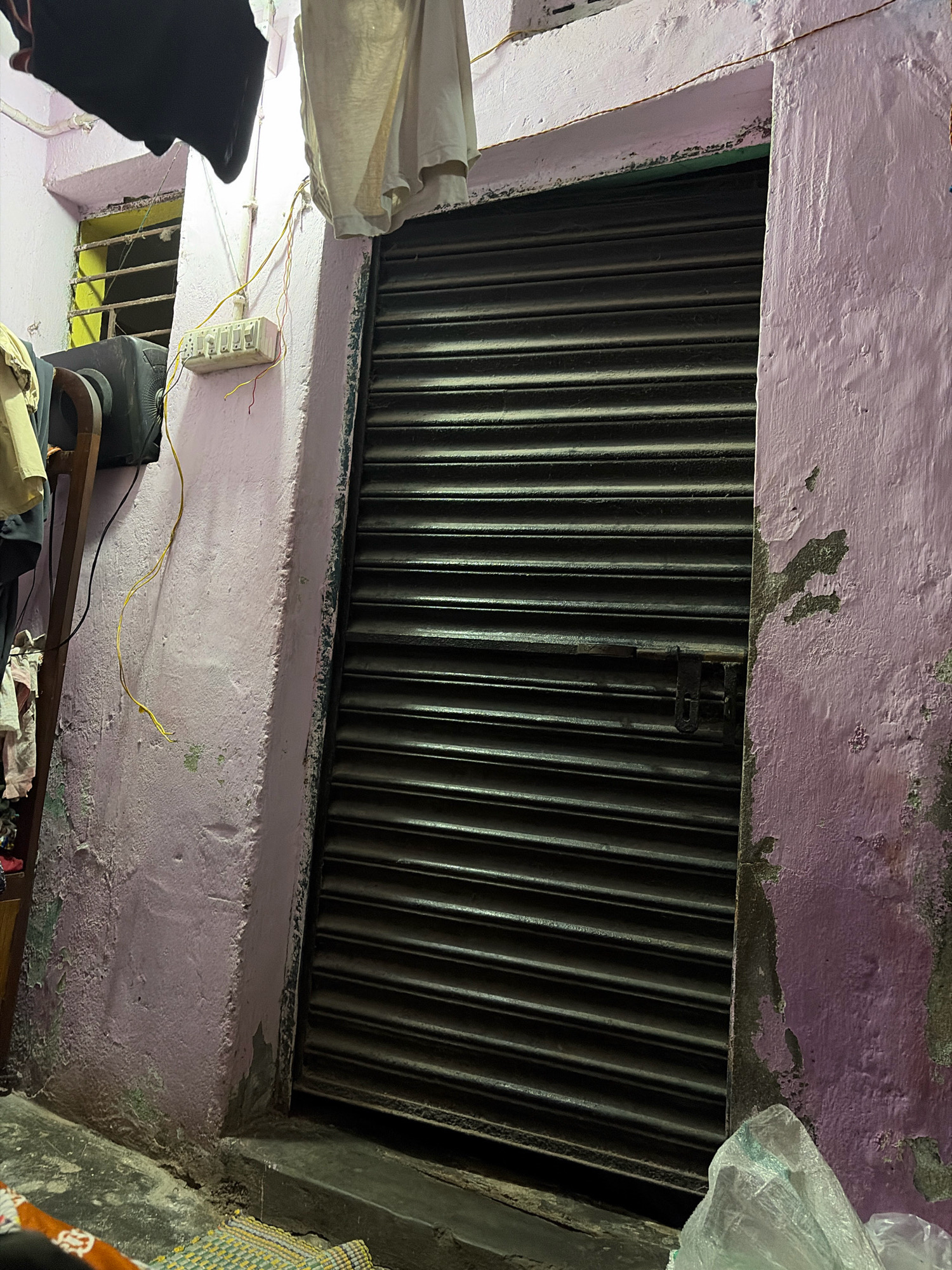 A narrow alleyway in Jamshedpur, India, is depicted, featuring a weathered pink concrete wall on the right, adjacent to a closed, dark gray metal roll-up door. The wall shows signs of peeling paint and patches of exposed concrete and some green discoloration. Above the door and to the left, laundry hangs from a clothesline, obscuring part of the upper frame. To the left of the roll-up door, a window with metal bars is visible, and to its left, a black television set rests on a wooden stand. A white electrical junction box with multiple switches is mounted on the pink wall, with yellow and red wires extending from it. The ground in the foreground is rough concrete, with a woven mat partially visible, and a plastic bag lies to the right. The lighting suggests it is either dusk or a dimly lit interior.