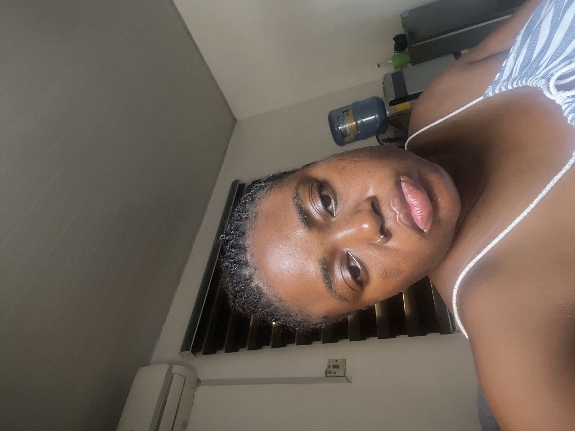 This is a close-up, portrait-style photograph of a young Black woman indoors, with the image rotated 90 degrees clockwise. She is looking directly at the camera, with her face partially visible. Her skin appears dewy and well-lit, with visible highlights on her forehead and cheek. She has a nose ring and appears to be wearing a light-colored tank top with thin straps, possibly striped.
In the background, a large blue water dispenser bottle is visible on a shelf, suggesting a home or office setting. There are also glimpses of blinds and an air conditioning unit, further indicating an indoor environment. The lighting suggests it might be evening or nighttime, as artificial light is predominantly used. The overall mood is intimate and casual. No text is clearly visible in the image. The context of Abuja, Nigeria, is noted but not visually evident in the provided image.