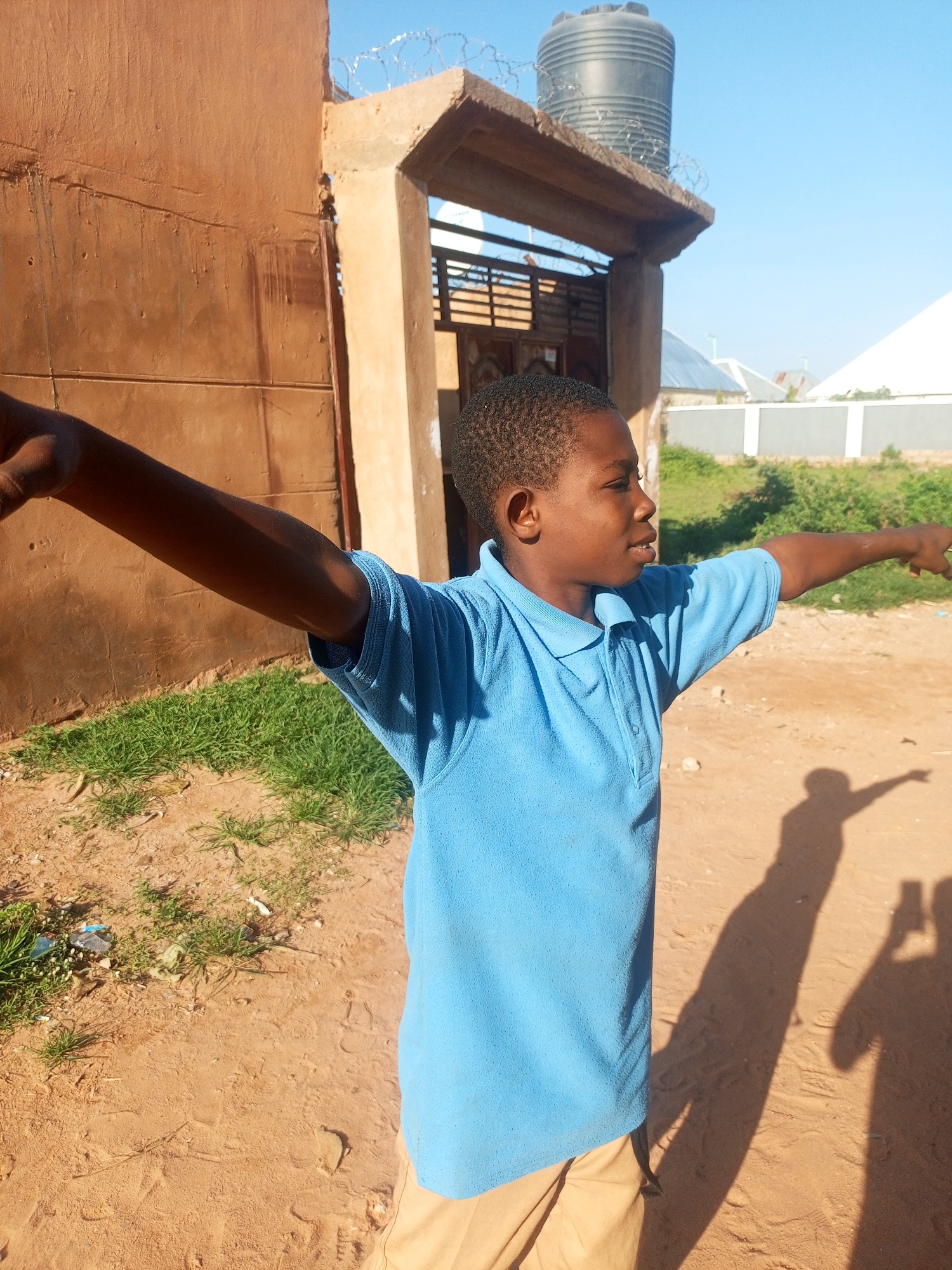 An outdoor, medium shot captures a young boy in Tempure, Nigeria. He is wearing a blue polo shirt and light brown shorts. His left arm is extended to the left, and his right arm is outstretched to the right, pointing. The boy is looking to his right, with a slight smile. The background consists of a sand-covered ground with some sparse green grass and a sandy-colored building with a concrete overhang. A black water tank is visible on top of the overhang, along with barbed wire. The sky is clear and blue. The boy's shadow and another shadow, possibly from the photographer, are cast on the ground.