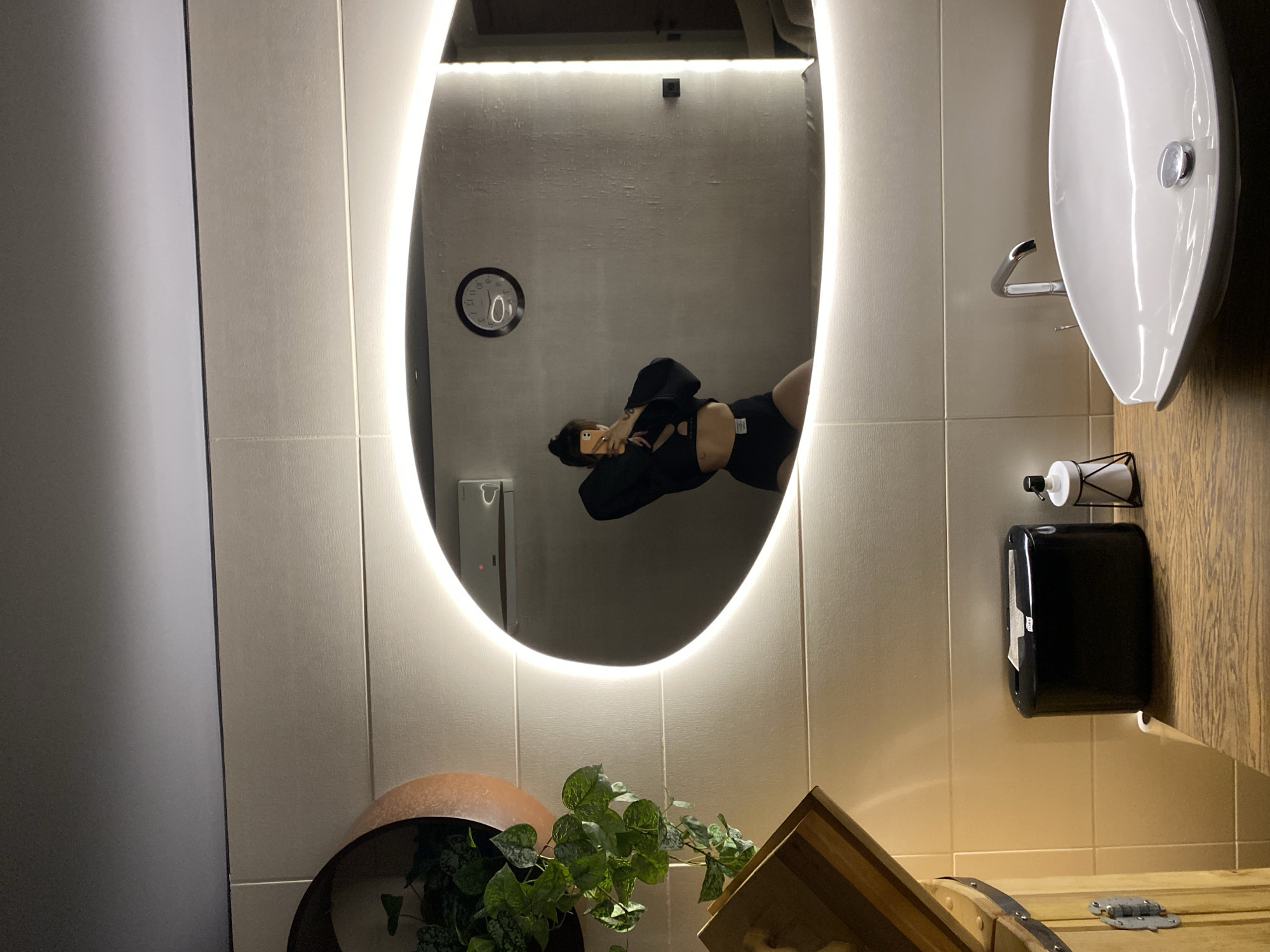 The image depicts a bathroom interior. The most prominent feature is a large, oval-shaped mirror with an illuminated edge, reflecting a person dressed in black activewear and holding a mobile phone in their hand. Reflected in the mirror is also a wall-mounted clock. Adjacent to the mirror, there is a white sink and faucet, alongside a black paper towel dispenser and soap dispenser. At the bottom of the frame, there are green plants. The tiles are beige. The bathroom counter is made of wood. The specific location in Votkinsk, Russia is not discernable from the image.