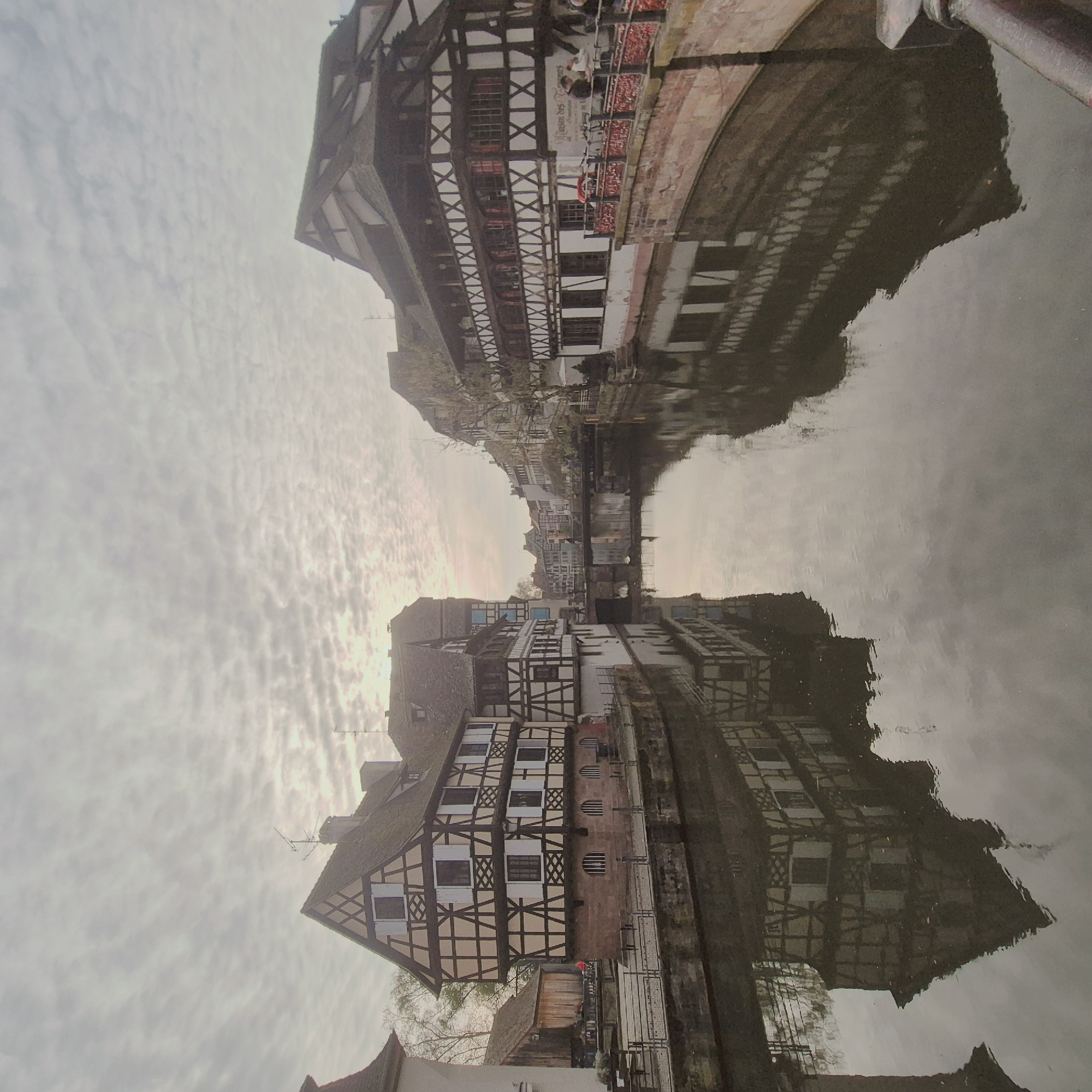 The image presents a detailed view of a canal in Petite France, Strasbourg, France, characterized by traditional half-timbered architecture. The sky is largely overcast with a pattern of altocumulus clouds, providing a diffuse, cool light. A calm canal occupies the lower and central portions of the frame, reflecting the sky and the adjacent buildings symmetrically.
On the left bank, a multi-story half-timbered house dominates the foreground, featuring a light-colored stucco or plaster infill contrasted by dark brown timber framing. It has a steeply pitched dark-tiled roof and multiple windows with white shutters. The ground floor of this building is constructed from a reddish-brown stone or brick foundation, with arched openings near the water level. Further back on the left, a simpler, brown wooden structure with a dark roof is partially visible. A narrow paved walkway with metal railings runs along the left bank.
Across the canal, on the right bank, another prominent multi-story half-timbered building, identified by the discernible inscription "Maison des Tanneurs," mirrors the architectural style with dark timber framing over a light facade and a dark-tiled roof. Its lower section is also composed of reddish stone. Several bicycles are parked against the building on this bank. A stone bridge with a curved balustrade adorned with numerous red and pink "love locks" extends from the right bank into the mid-ground.
Several individuals are visible. On the right, a person in a dark jacket stands near the bridge railing, and another person in a red top is visible near the bicycles. Further down the canal, a few indistinct figures are walking across a pedestrian bridge. At least two additional bridges span the canal in the distance. Bare deciduous trees are scattered along both banks. Television or radio antennas are visible on the rooftops of several buildings on both sides of the canal.