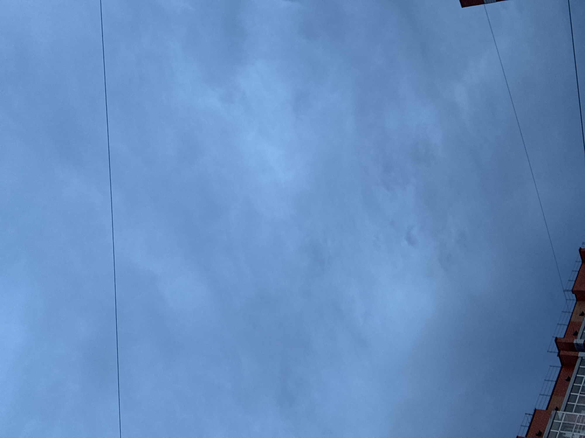 The image depicts a cloudy, overcast sky dominating the frame, with a subtle gradient of deep blue to lighter hues. A thin, dark horizontal line, likely a wire, bisects the sky roughly in the upper third of the image. In the lower portion of the image, the top of a brick building is visible. The building features red brickwork, a white-framed window structure, and a metal railing along its edge. The overall scene suggests an outdoor urban environment, possibly during twilight or a heavily clouded day. The location is identified as Markova, Russia. There are no people or specific actions observed in the image.