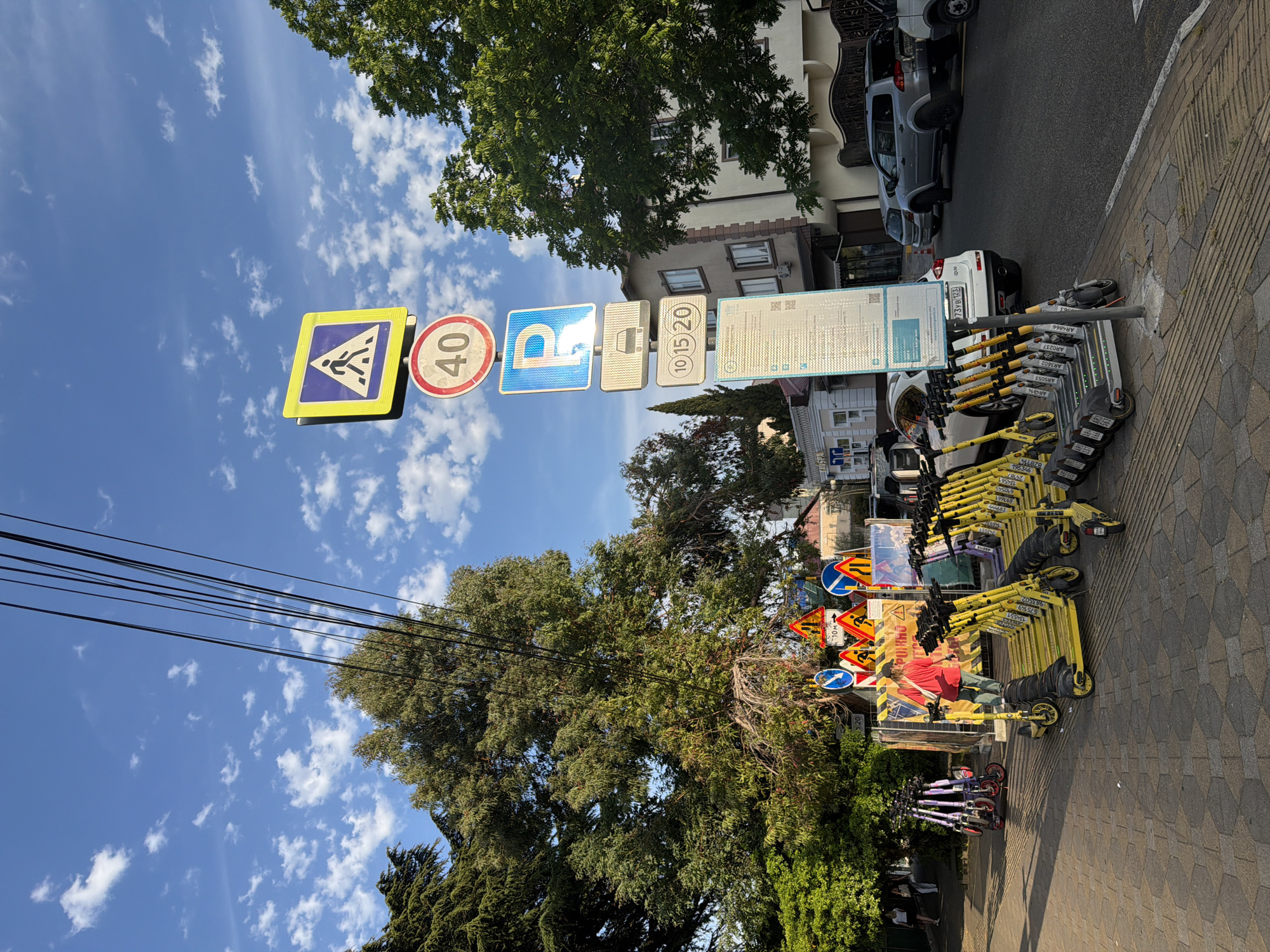 The image captures a sunny street scene in Sochi, Russia, on a day with a bright blue sky dotted with white clouds. A prominent cluster of street signs is mounted on a pole in the foreground, featuring a pedestrian crossing warning, a speed limit of 40, a parking symbol, a lane restriction for vehicles, and time restrictions of 10, 15, and 20 minutes. Below these, a large information board is attached to the pole.
To the left of the signs, a row of yellow electric scooters is parked along a paved sidewalk. A woman in a red shirt and green pants is standing near the scooters, facing them. Further left, more scooters, this time purple, are propped against a railing, with two people walking in the background.
The sidewalk is paved with hexagonal tiles. Behind the scooters and the people, a street lined with cars and a white SUV is visible. Trees, including a large, leafy evergreen, frame the scene. The background shows buildings with windows and a fence. Overhead, power lines are strung across the sky. The atmosphere appears calm and pedestrian-friendly, with evidence of shared mobility options.