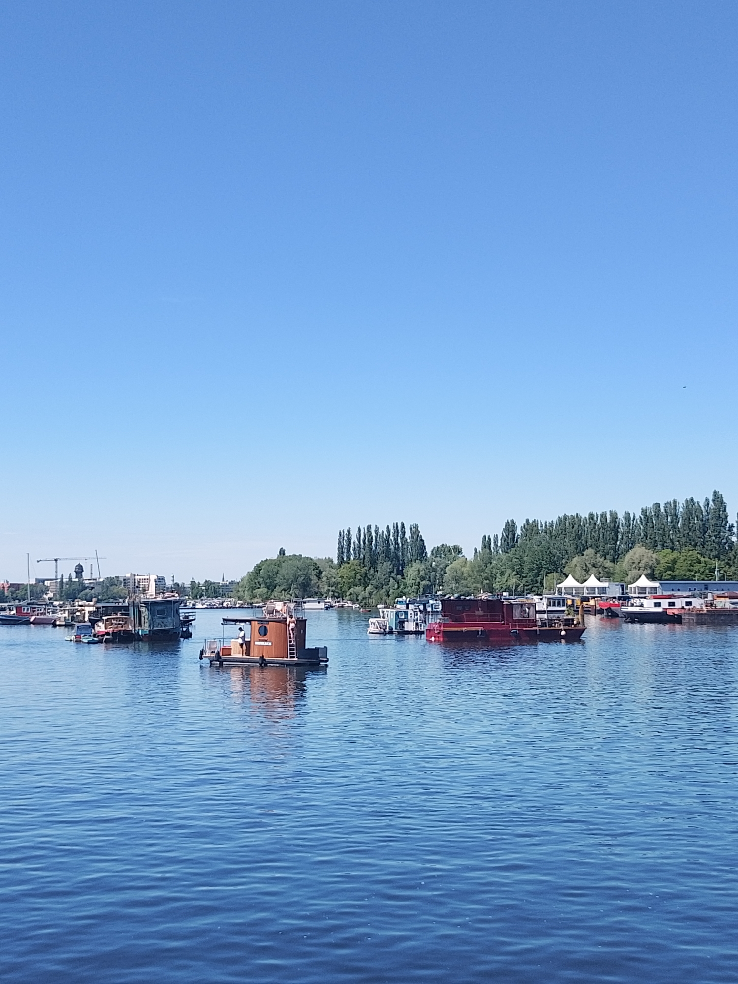 The image shows a body of water with several boats and houseboats anchored or moored on it. The water appears calm with slight ripples. The boats vary in size and color, including red, brown, and gray. On the shoreline, there are trees and structures, including white tents, suggesting a recreational area or waterfront development. In the background, a cityscape is visible with buildings and a crane. The sky is clear and blue, indicating a sunny day. The location may be a river or large lake in a city. The city is Unknown, Unknown.
