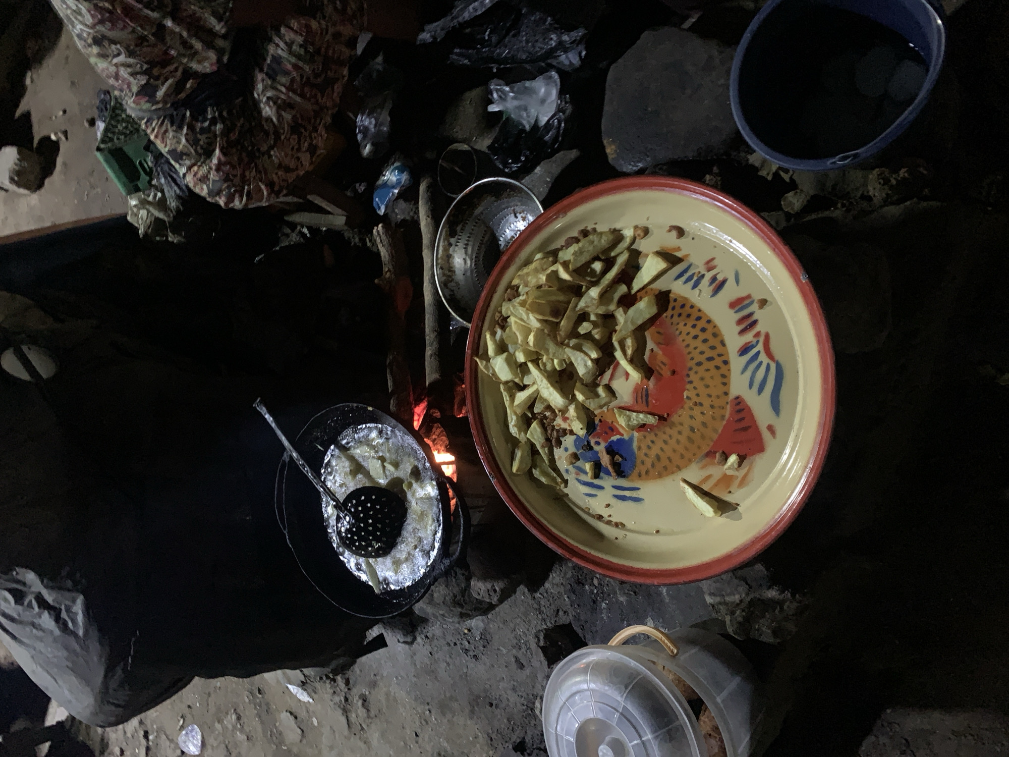 A ground-level shot shows a cooking setup outdoors, likely in a rural or semi-rural environment in the city of Rafin Par, Nigeria. A plate with cooked french fries is the primary focus. The plate is ceramic with a red rim and a fish design in the center. To the left, a black pot sits over a fire, with more french fries visible, and a slotted spoon rests in the pot. A metal strainer is nearby. A clear plastic container and a blue bucket are also visible. The background is mostly dark, with the light concentrated on the cooking area. There is a person or people out of view. The cooking area consists of rocks used as a foundation.