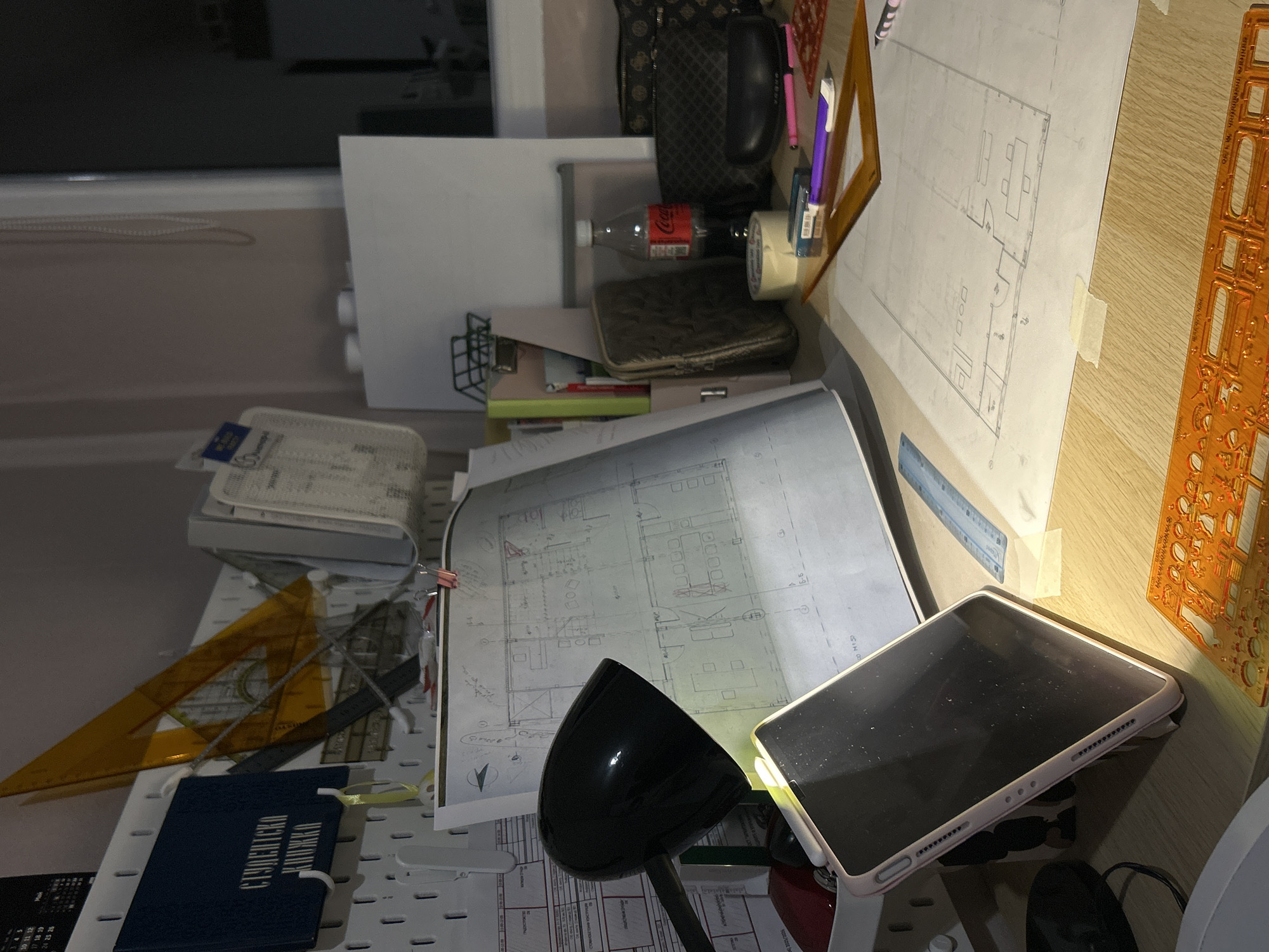 The image shows an untidy desk in what appears to be a home or office setting. The desk is covered with various architectural and office supplies, including drafting paper with floor plans, a black desk lamp, a tablet, triangles, a ruler, a student's notebook, a stencil, a Coca-Cola bottle, and personal belongings like a purse. The lighting is uneven, with some areas in shadow. The photo was not necessarily taken in Sofia, Bulgaria, and no identifiable location is visible in the image.