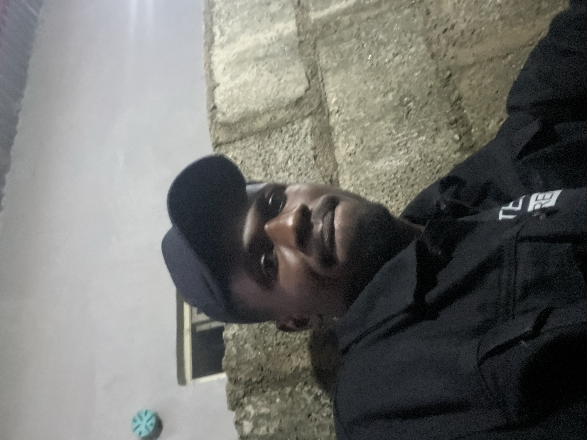 One adult male is captured in a close-up, slightly low-angle shot within an unspecified location in Unknown, Unknown. The subject has a dark complexion, a beard, and wears a black baseball cap. He is dressed in a dark, likely black, collared jacket or shirt, which displays partial white text, "TE" above "RE," on the left chest area. His gaze is directed towards the camera with a neutral to slightly smirking expression. The background features various wall textures: in the upper left, a plain light grey wall holds a circular light-blue decorative object with a star-like pattern. Below this, a dark rectangular opening, possibly a window or vent, is visible. To the right and behind the subject, a wall constructed from greyish-brown concrete blocks with visible mortar lines forms the primary background. The overall lighting conditions are dim, suggesting an evening or poorly lit indoor/outdoor environment. No specific actions or interactions are discernible beyond the subject posing for the image.