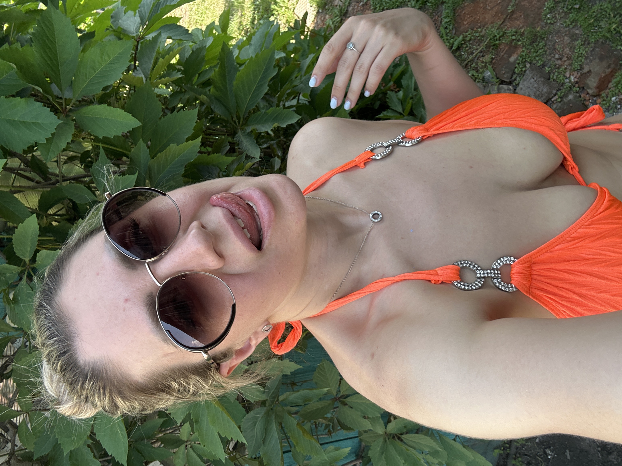 A woman in a bright orange bikini and sunglasses is posing for a selfie outdoors. She has her tongue sticking out and is looking at the camera. The background is lush green foliage and a wooden fence. The image is taken in Novoaltaysk, Russia, during the daytime.