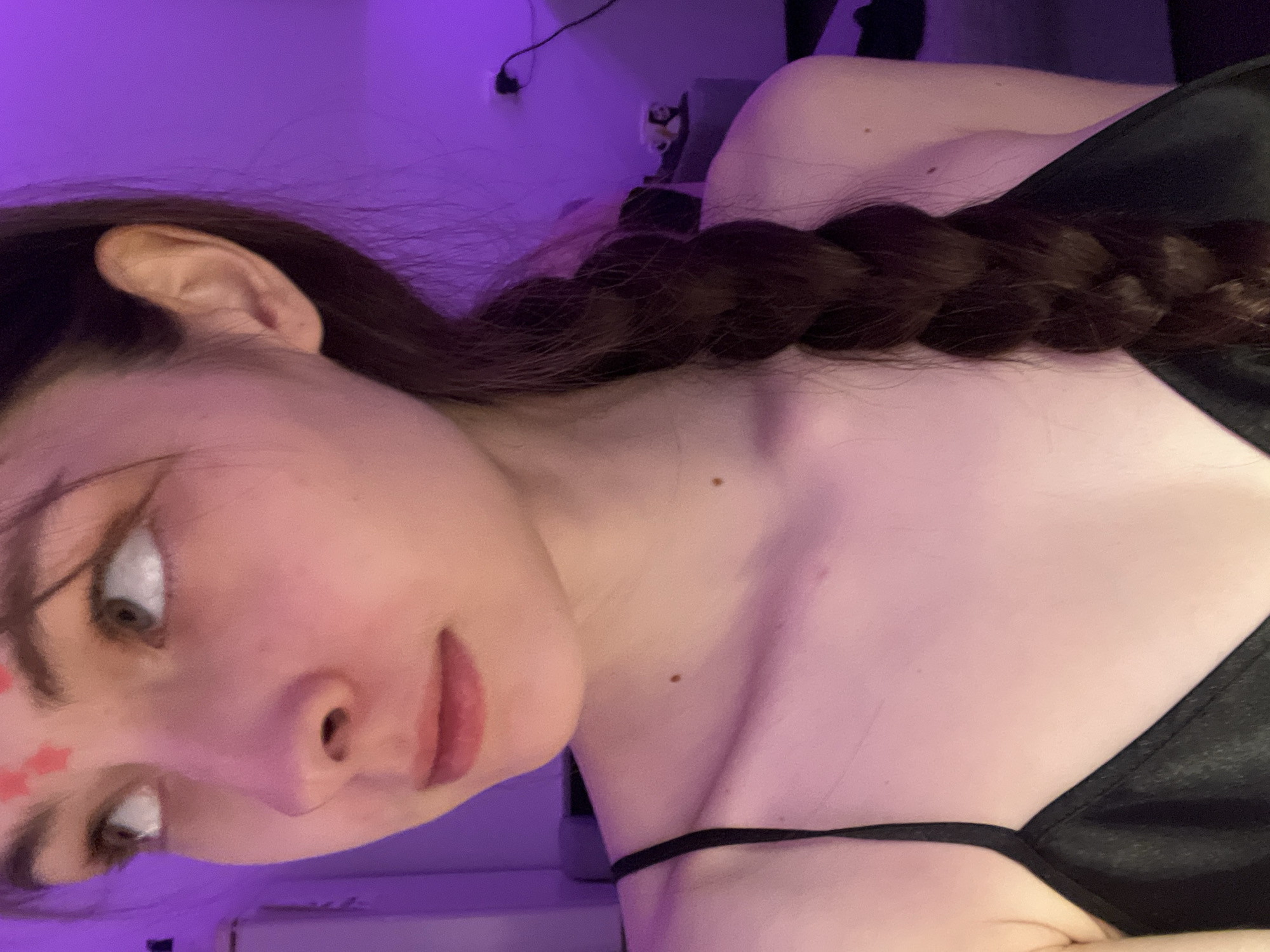 A young woman is depicted in a close-up shot, angled slightly, revealing her face and upper body. She has fair skin, brown hair styled in a braid, and a red star sticker on her forehead. She's wearing a black garment with thin straps. The background is a purple hue, and objects like a wall outlet and a figure are visible in the blurred area. There's no contextual information suggesting this image was taken in Saint Petersburg, Russia.