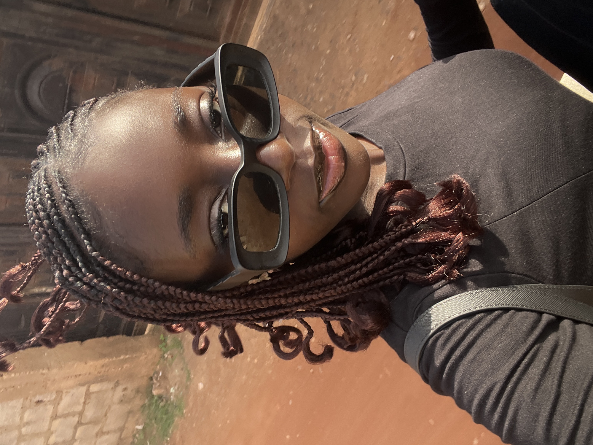 A medium, angled shot captures a brown-skinned woman in Abuja, Nigeria. She wears a black t-shirt and sunglasses, with reddish-brown braided hair. A strap crosses her shoulder. The backdrop features a worn brick wall and a reddish-brown textured ground.